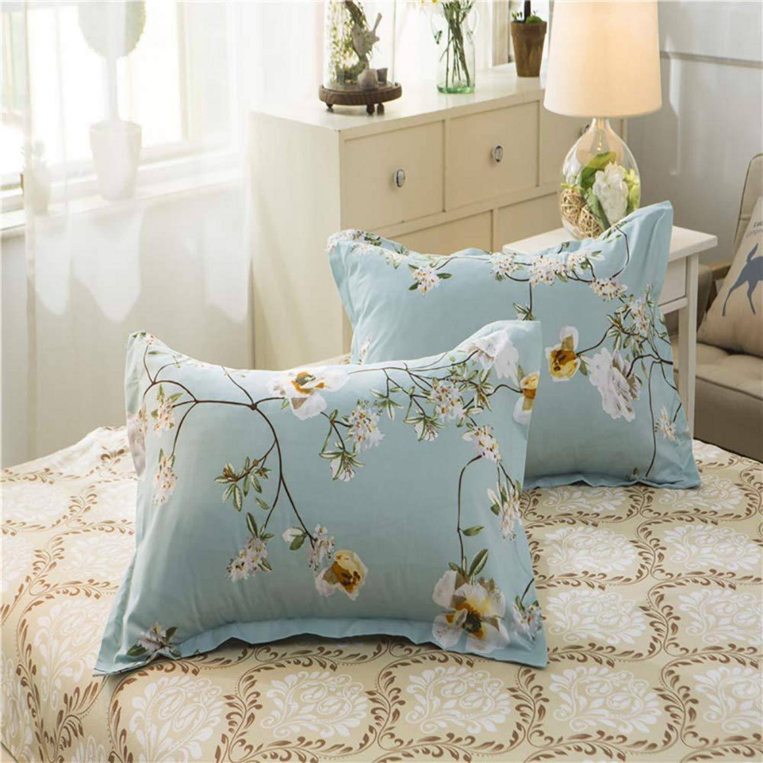 Zwei hellblaue Kissenbezüge mit Blumenmuster auf einem Bett in einem hellen Schlafzimmer, mit einer cremefarbenen gemusterten Bettdecke und einer Nachttischlampe.