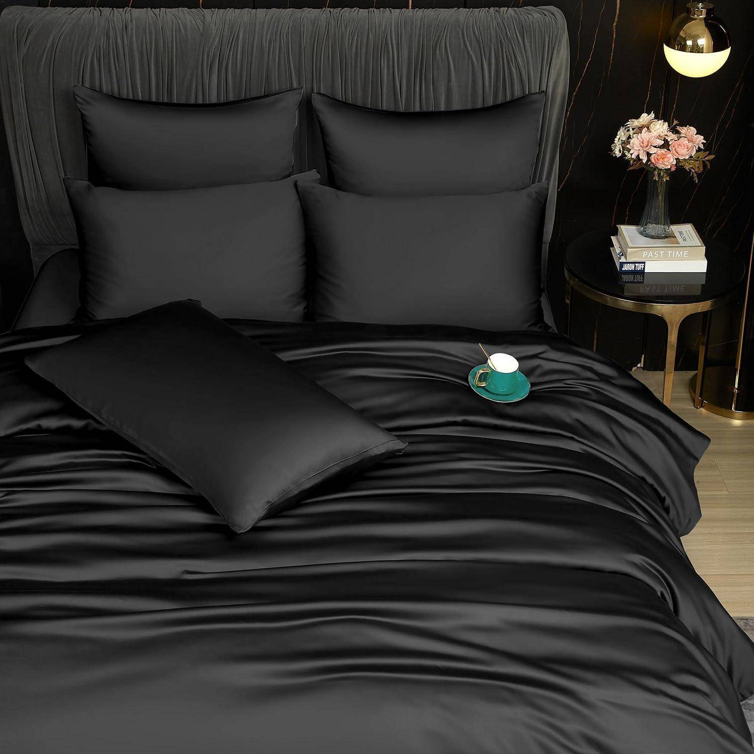 Schwarze Satin-Bettwäscheset mit Kissenbezügen und einem Dekokissen auf einem ordentlich gemachten Bett neben einer kleinen Lampe und einem Nachttisch mit Blumen.