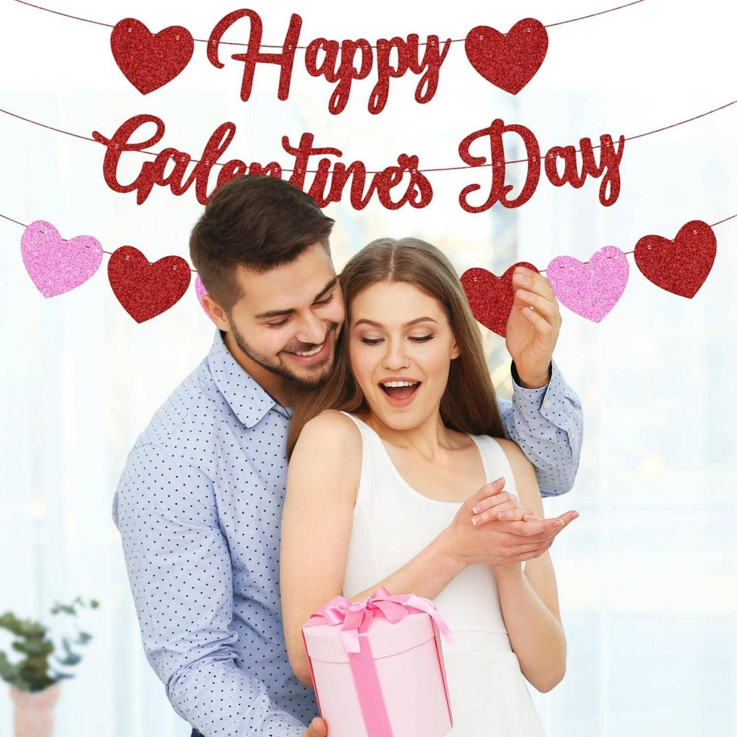 Ein lächelndes Paar, das sich auf einer Valentinstagsfeier umarmt, wobei der Mann der Frau eine rosa Geschenkbox unter einem „Happy Valentines Day