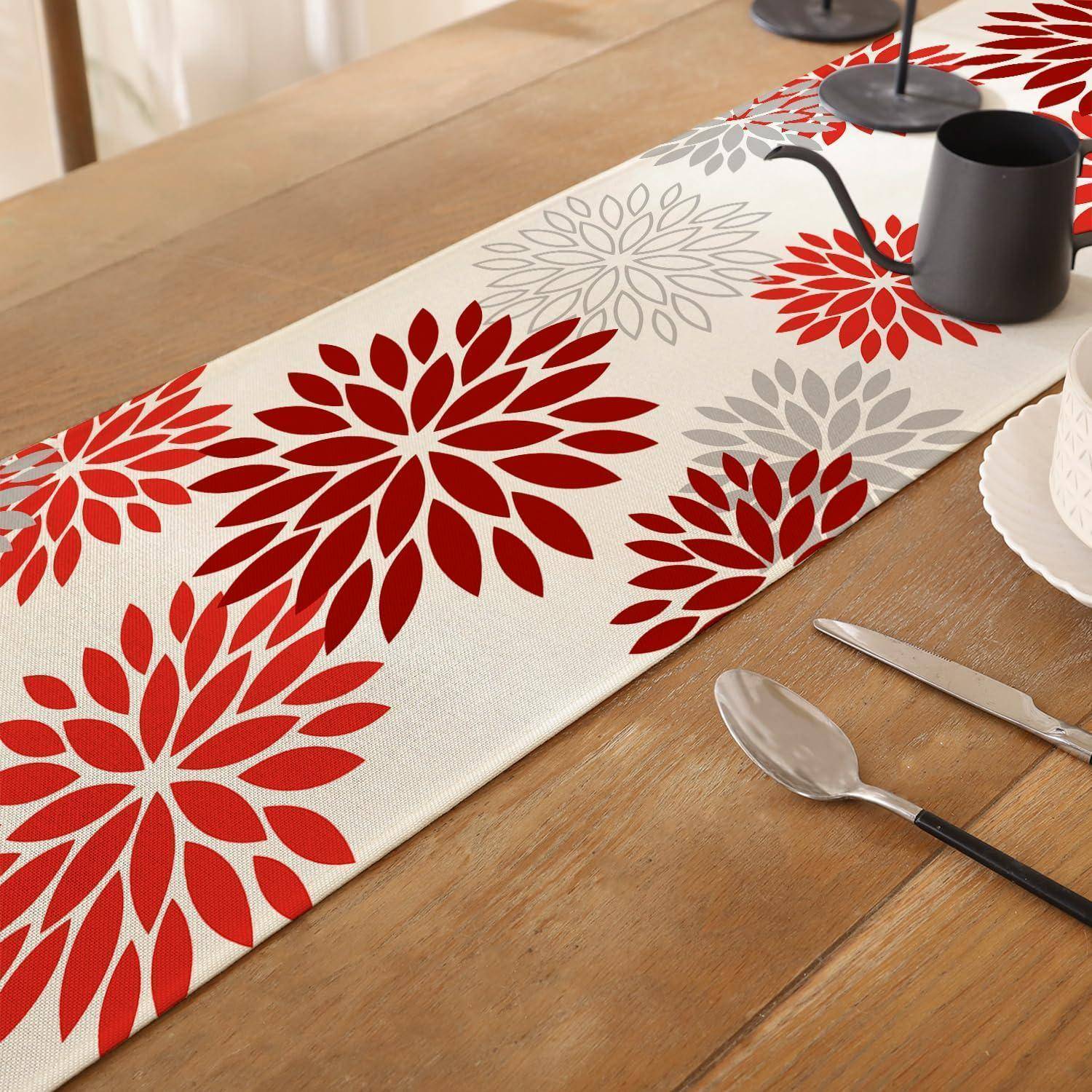 Ein hölzerner Esstisch mit einem weißen Läufer, der mit roten und grauen Blumenmustern bedeckt ist, eingedeckt mit einer schwarzen Tasse, Besteck und Tellern.