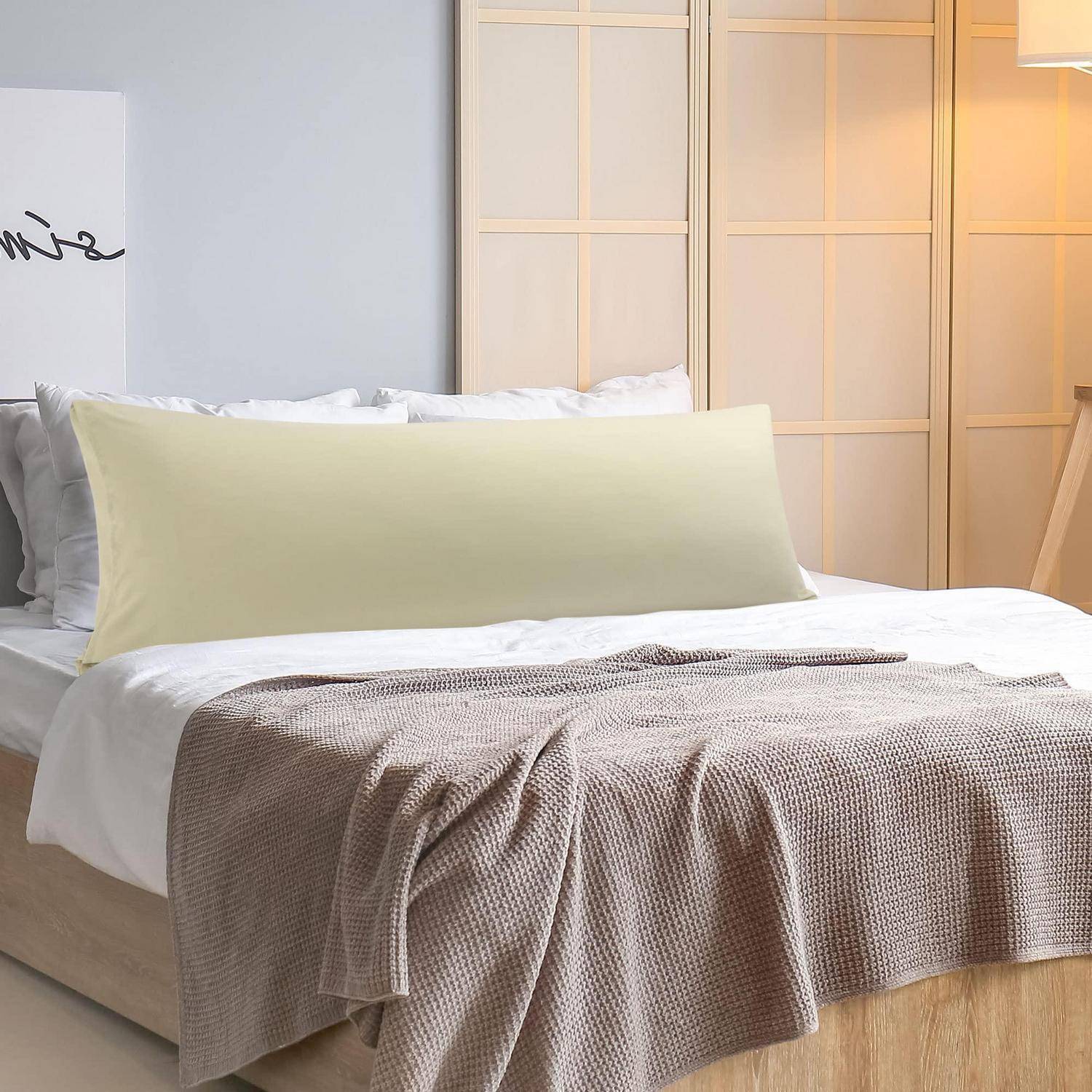 Ein ordentlich gemachtes Bett mit weißer Bettwäsche, einem hellgrünen Seitenschläferkissen und einer beigen Wurfdecke in einem sanft beleuchteten Schlafzimmer.