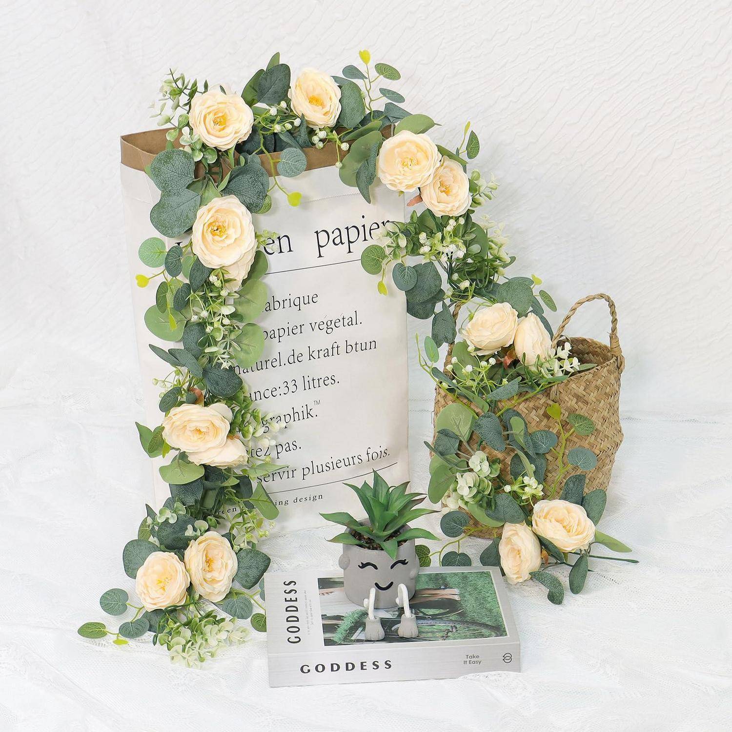 Dekorative Blumengestaltung mit cremefarbenen Rosen und Eukalyptus, drapiert um eine Papiertüte, neben einem geflochtenen Korb und einer kleinen Topfpflanze mit Sukkulenten auf weißem Hintergrund.
