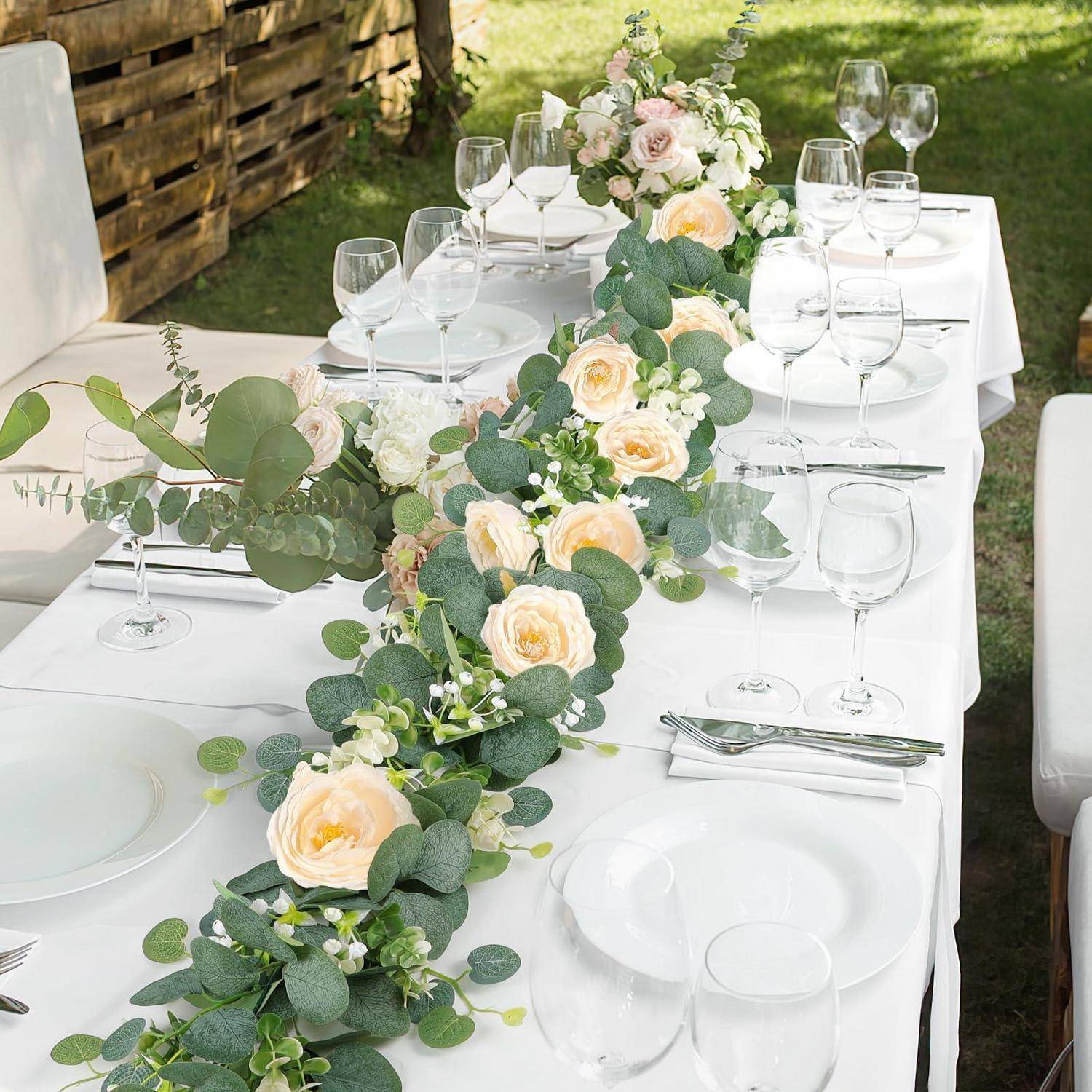 Eleganter Außenessplatz mit weißem Tischtuch, Gläsern, Tellern und einer langen Blumendeko aus cremefarbenen Rosen und Grün.
