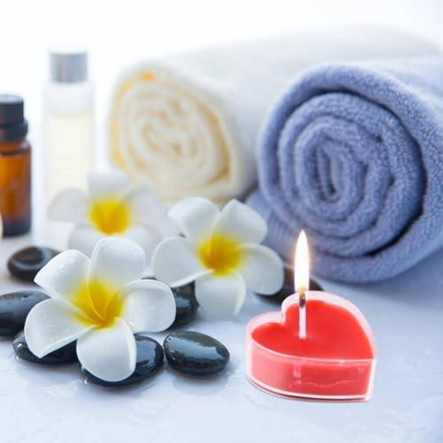 Spa-Stillleben mit aufgerollten Handtüchern, weißen Plumeria-Blüten, Massagesteinen, Ölen und einer brennenden herzförmigen roten Kerze.