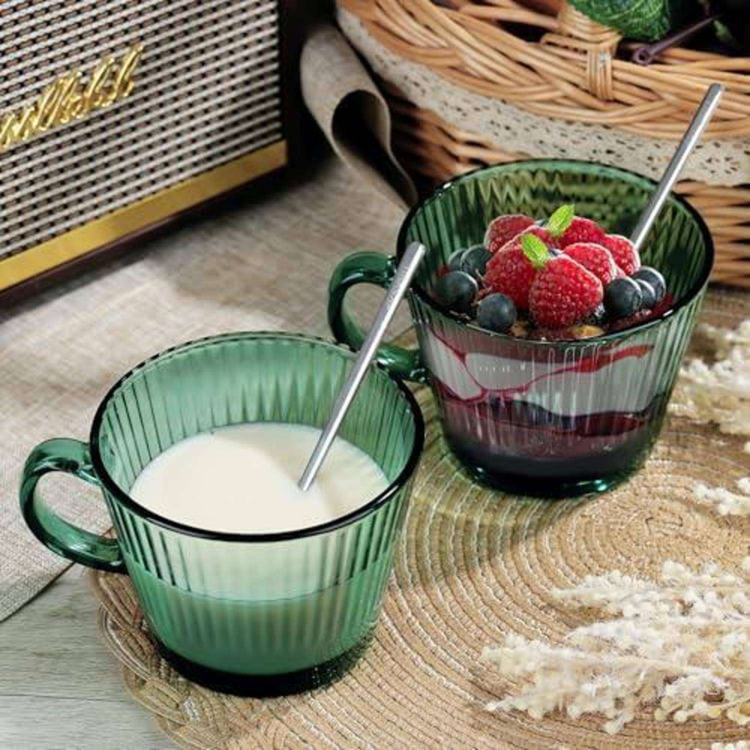 Zwei grüne Glastassen auf einer Flechtmatte, eine mit Milch und Löffel, die andere mit geschichtetem Joghurt, garniert mit Himbeeren, Blaubeeren und Minze.
