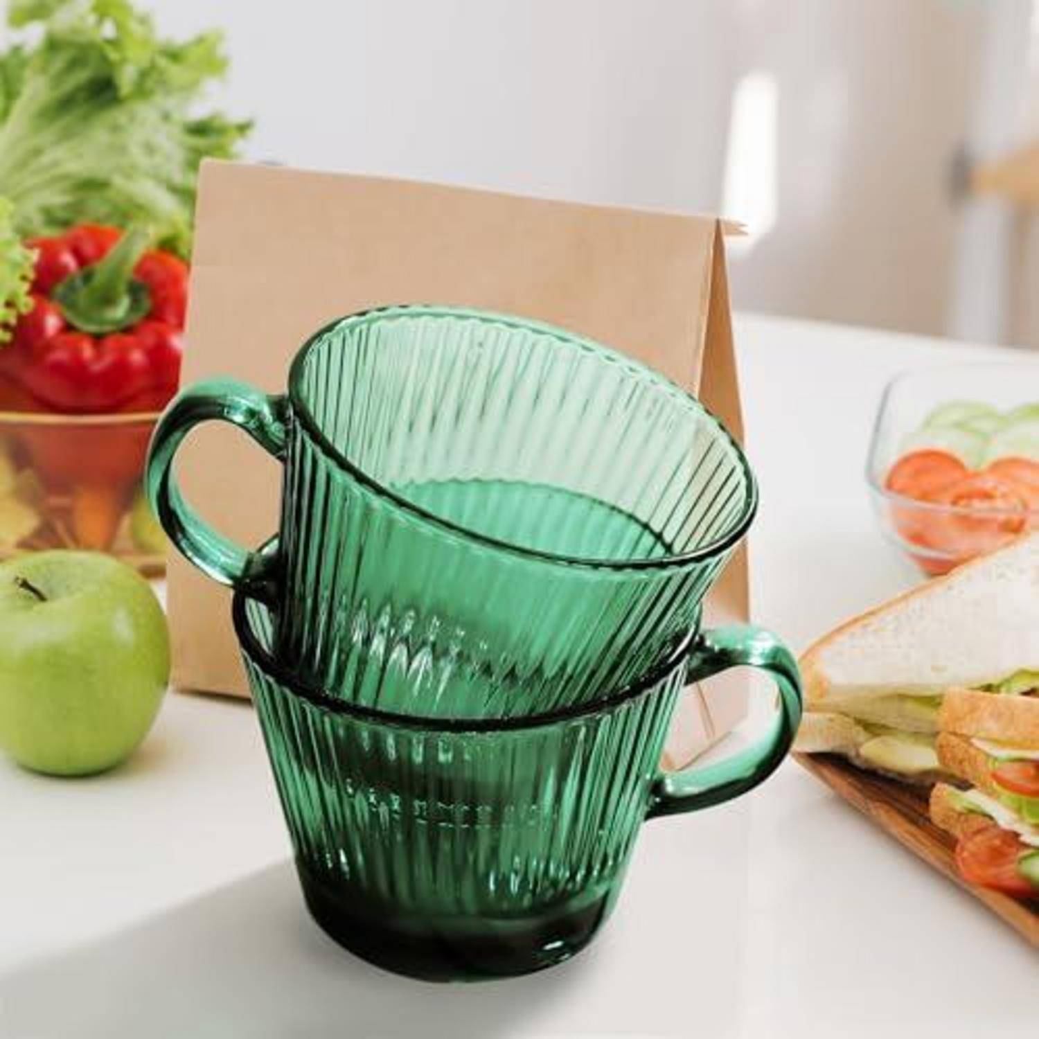 Zwei übereinander gestapelte grüne Glastassen auf einem Tisch mit einem Lunch-Setup, einschließlich Obst, Sandwich, Tomaten und einer Pappschachtel im Hintergrund.