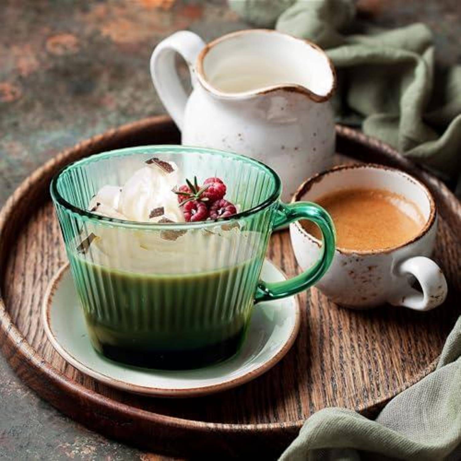Grüne Glastasse mit Matcha-Dessert, garniert mit Schlagsahne und Beeren, serviert auf einem Holztablett mit einer kleinen Kaffeetasse und einem Sahnekännchen.