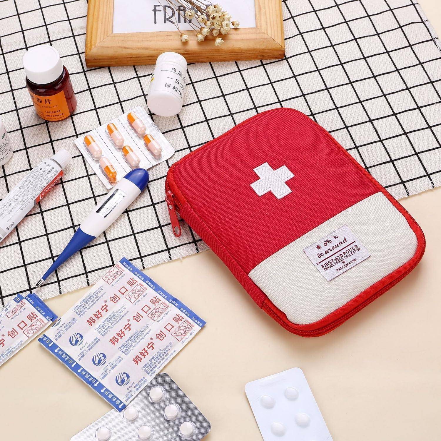 Rote Erste-Hilfe-Tasche mit weißem Kreuz, umgeben von verschiedenen Medikamenten, Pillen, Salbe und einem Thermometer auf einem karierten Tuch.