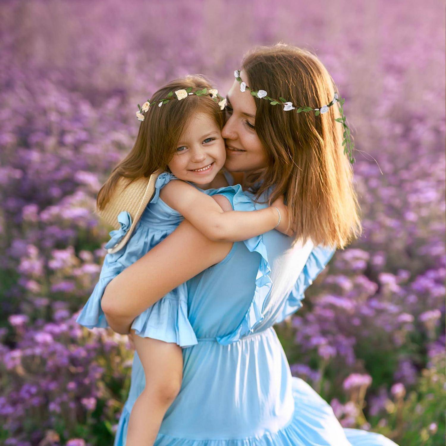 Mutter hält und umarmt ihre lächelnde junge Tochter in einem violetten Blütenfeld, beide tragen hellblaue Kleider und Blumenkränze.