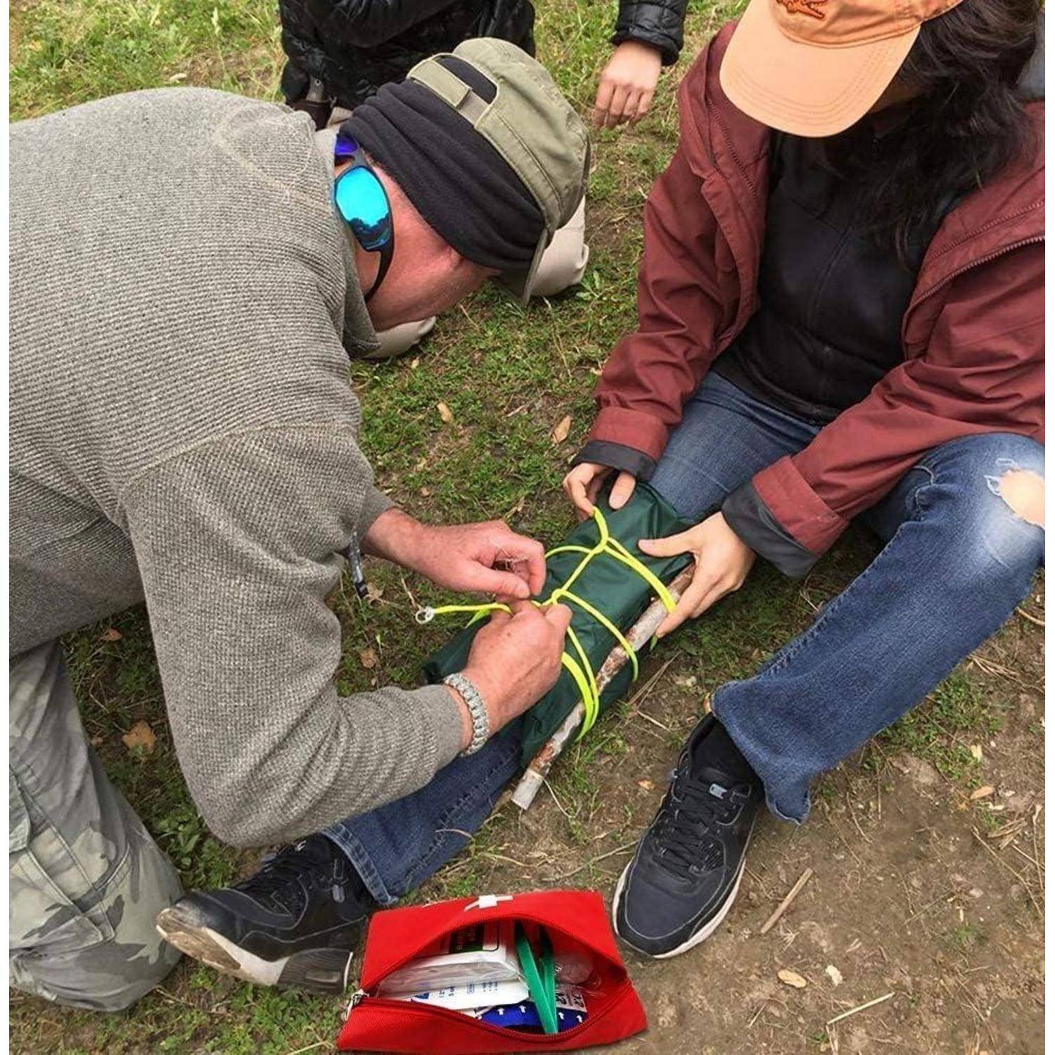 Menschen im Freien, die Erste Hilfe üben: Eine Person zieht ein Tourniquet um das Unterschenkel einer anderen Person fest, während andere in der Nähe zusehen, mit einem roten Erste-Hilfe-Kasten auf dem Boden.
