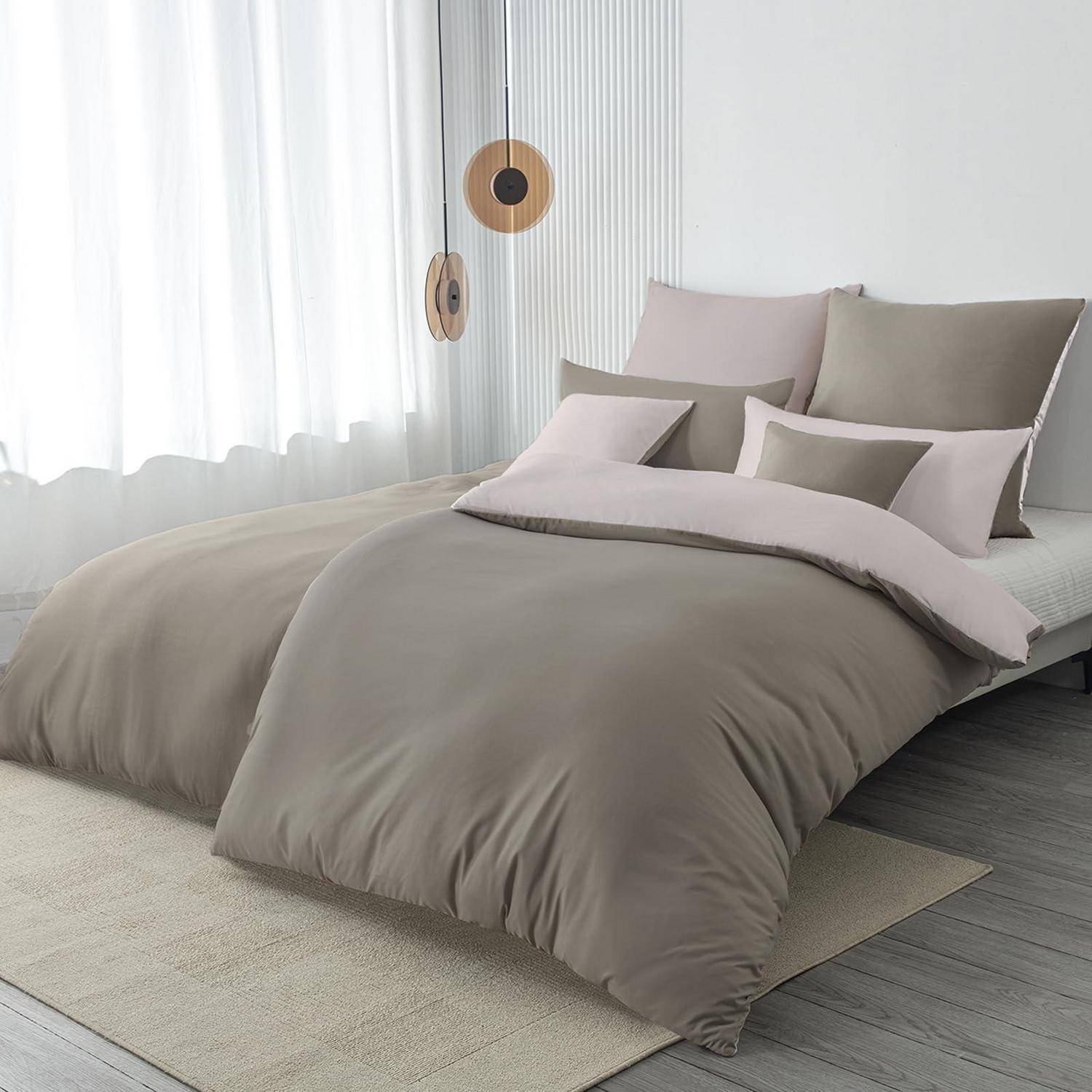 Ein ordentlich gemachtes Bett mit taupe- und beigefarbener Bettwäsche und Kissen in einem hellen, minimalistischen Schlafzimmer neben durchscheinenden Vorhängen und hängenden Pendelleuchten.