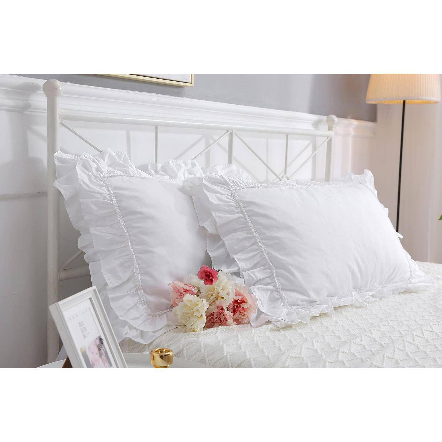 Weißes Bett mit gerüschten Kissen, ein kleiner rosa-weißer Blumenstrauß und eine Nachttischlampe in einem hellen Schlafzimmer.