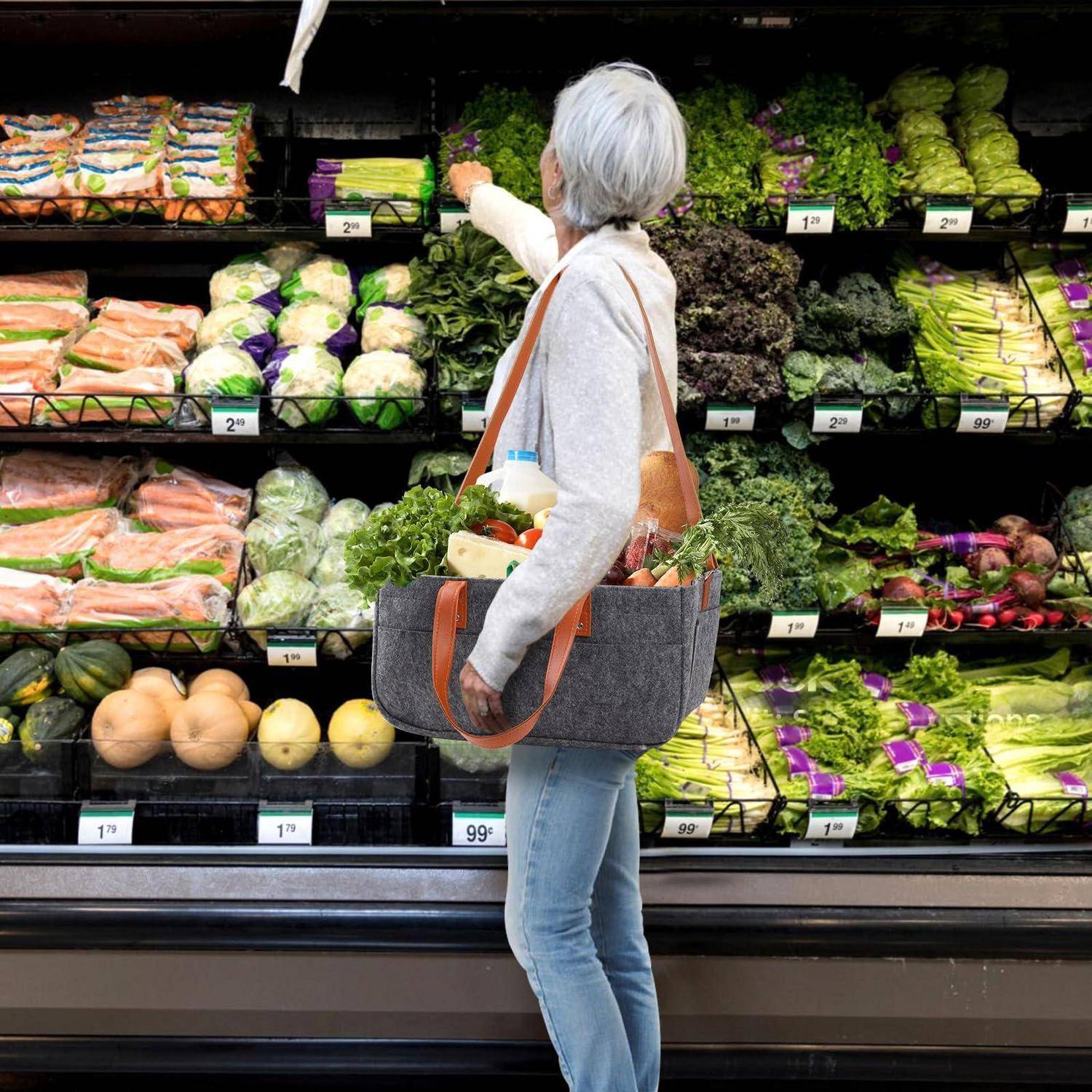 Frau beim Einkaufen in der Obst- und Gemüseabteilung eines Supermarkts, die nach Blattgemüse greift, während sie eine wiederverwendbare Tasche trägt und einen Einkaufswagen voller Gemüse schiebt.