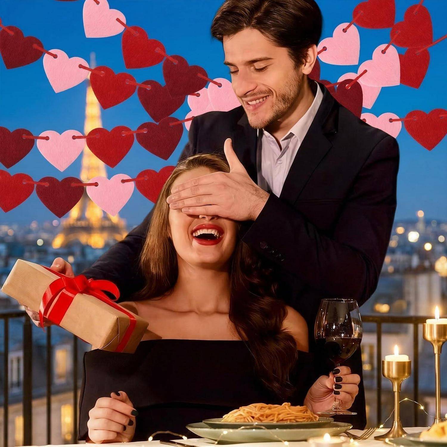 Ein Mann bedeckt einer Frau die Augen, während sie lächelt und ein Geschenk, Wein und Pasta bei einem romantischen Abendessen bei Kerzenlicht mit Herzdekorationen hält.