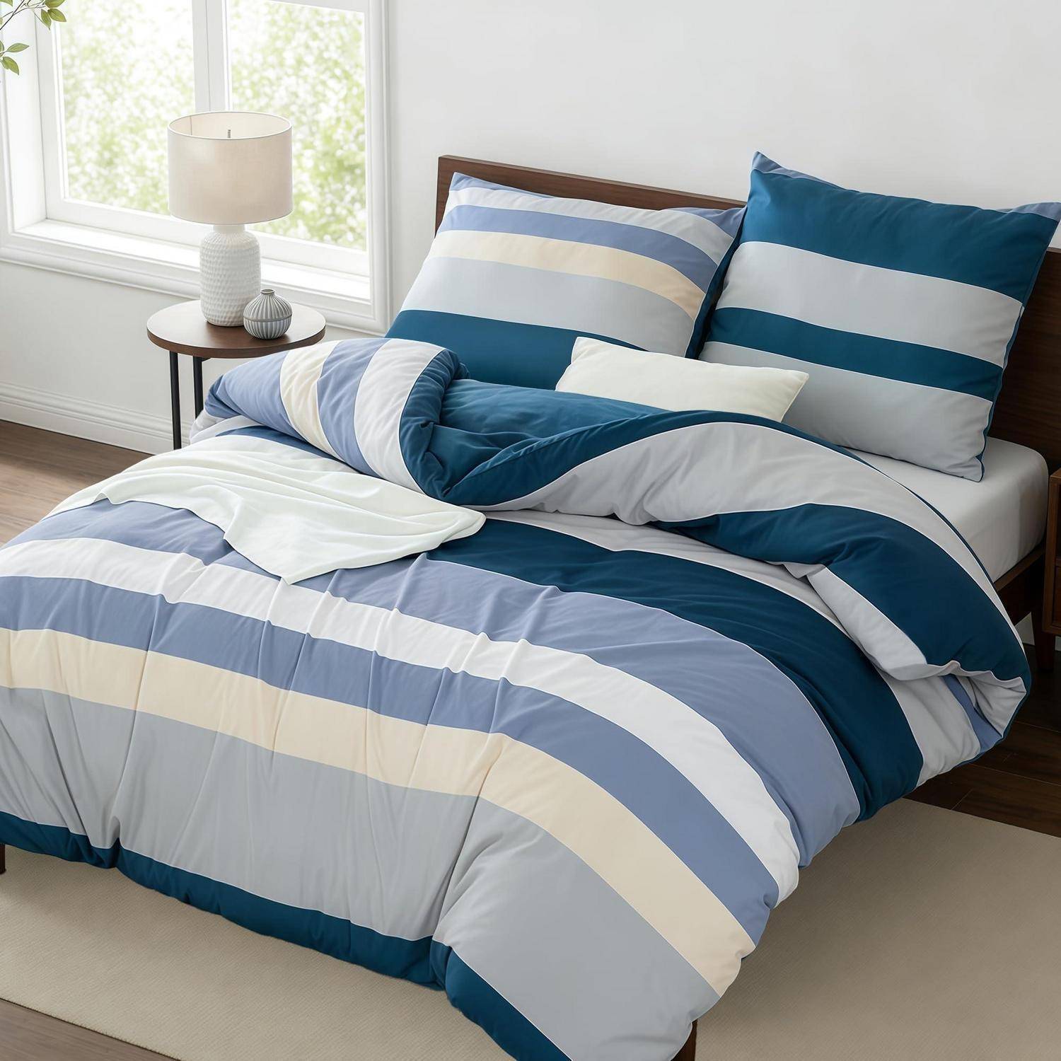 Ordentlich gemachtes Bett mit blau-weiß-cremefarbener gestreifter Steppdecke und Kissen in einem hellen Schlafzimmer neben einem Fenster und einer Lampe.