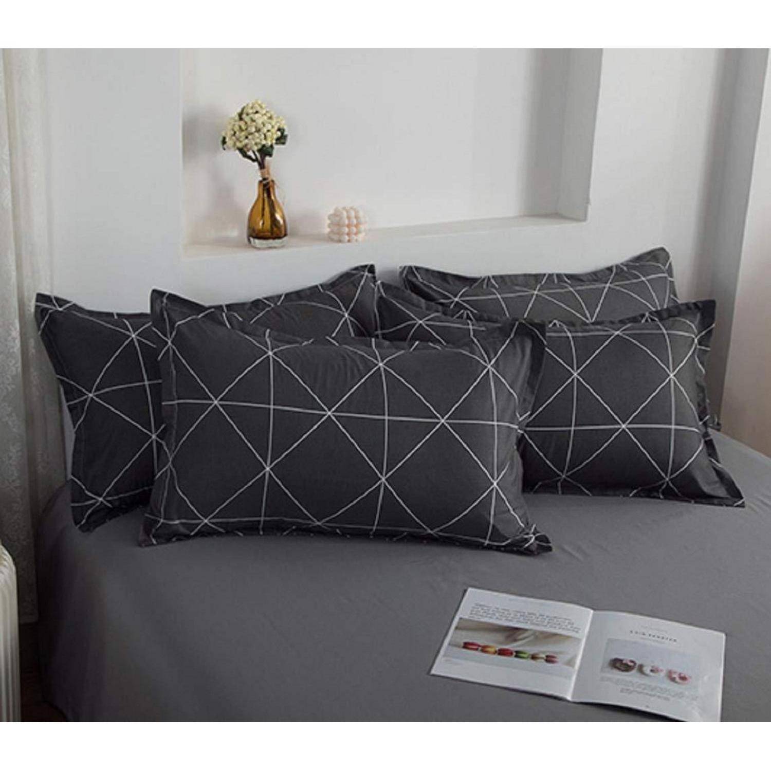 Bett mit grauer Bettwäsche und schwarzen geometrischen Kissen, mit einer offenen Zeitschrift auf der Decke und einer kleinen Vase auf einem Wandregal dahinter.