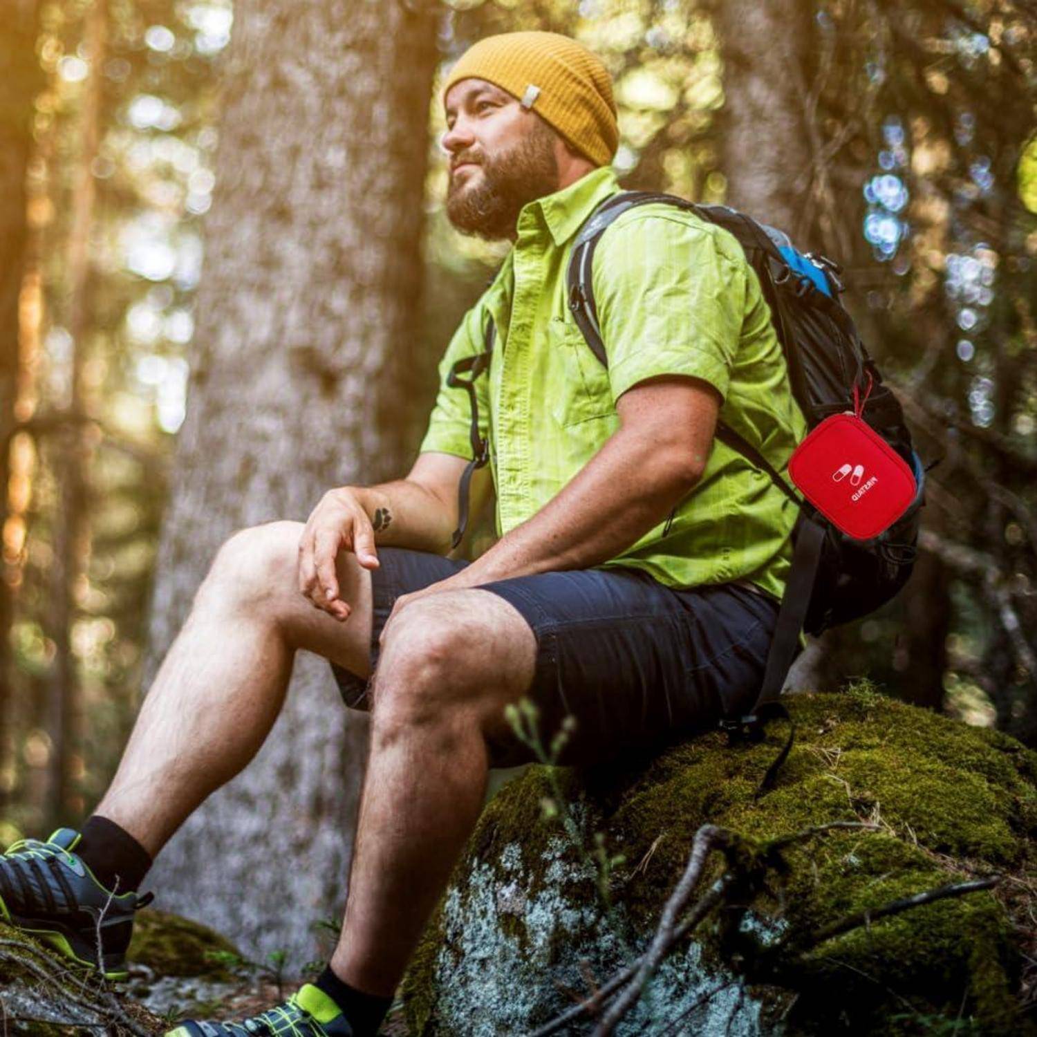 Bärtiger Wanderer in einer gelben Mütze und grünem Hemd, der auf einem moosbedeckten Stein im Wald ruht, mit einem Rucksack, an dem ein rotes Erste-Hilfe-Set befestigt ist.