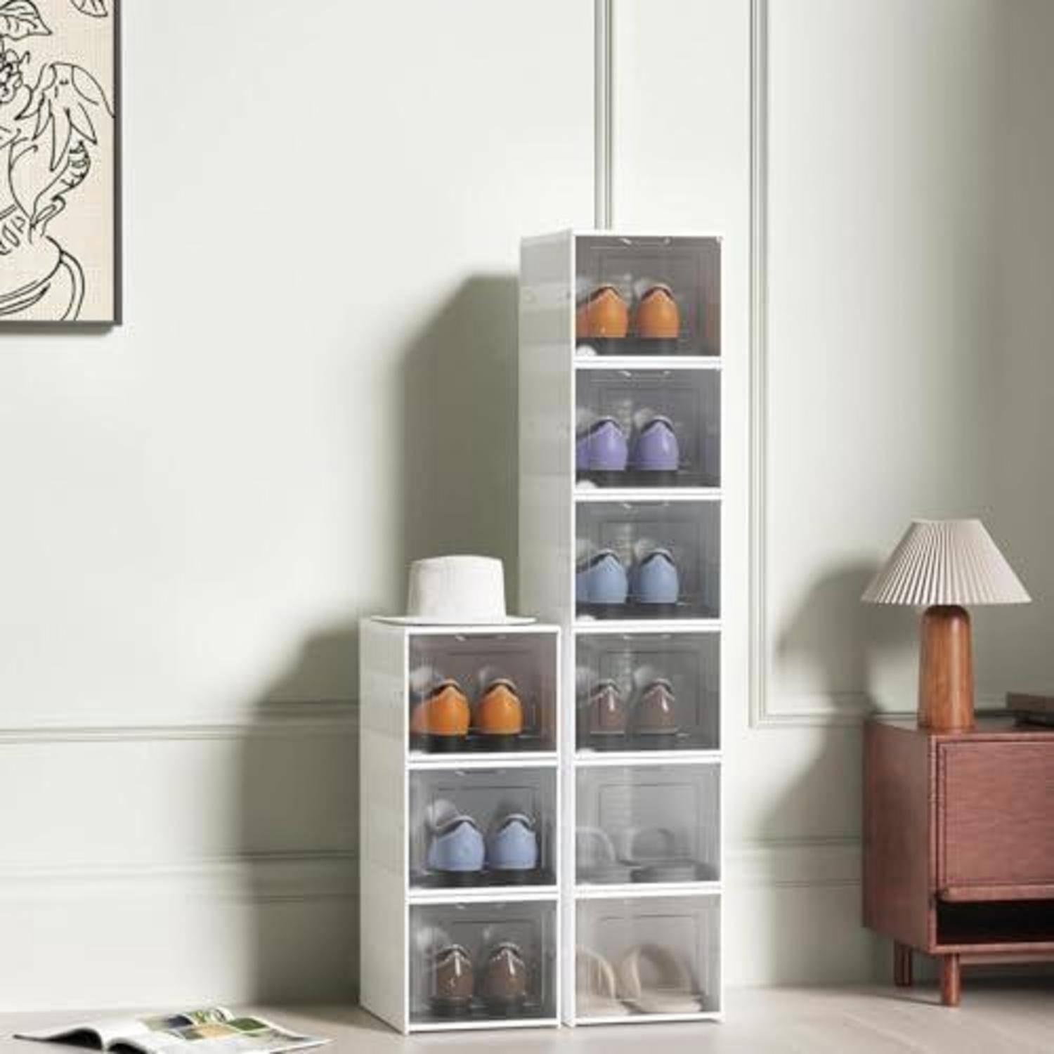 Gestapelte transparente Schuhaufbewahrungskisten mit Schuhpaaren in einem ordentlichen Zimmer neben einem Holzschrank und einer Tischlampe.