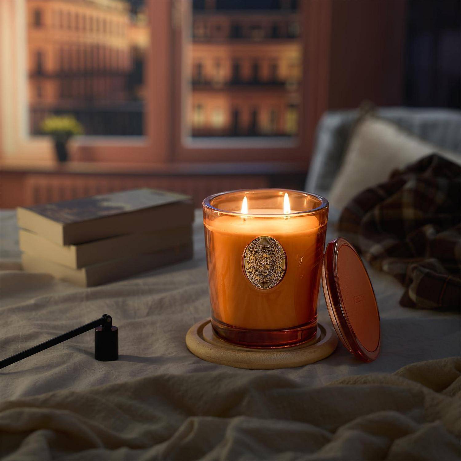 Brennende orangefarbene Kerze in einem Glaskrug auf einer Nachttischfläche, mit Büchern und einem Kissen im Hintergrund, die eine gemütliche Abendszene schaffen