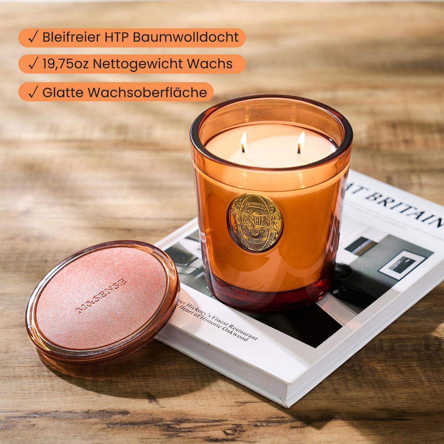Bernsteinfarbenes Kerzenglaskerzenglas mit zwei brennenden Dochten neben seinem passenden Deckel auf einer Zeitschrift; deutscher Text hebt Baumwolldocht, 19,75 oz Wachs und glatte Wachsoberfläche hervor.