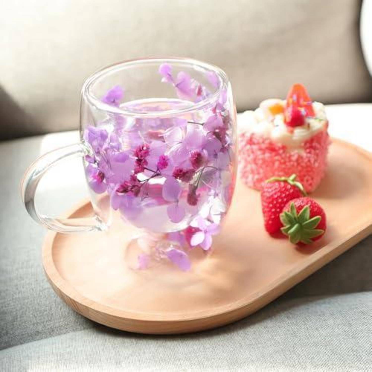 Glaskrug gefüllt mit violetten Blumen auf einem Holztablett neben einem Dessert mit Erdbeeren und frischen Erdbeeren auf einem Sofa.