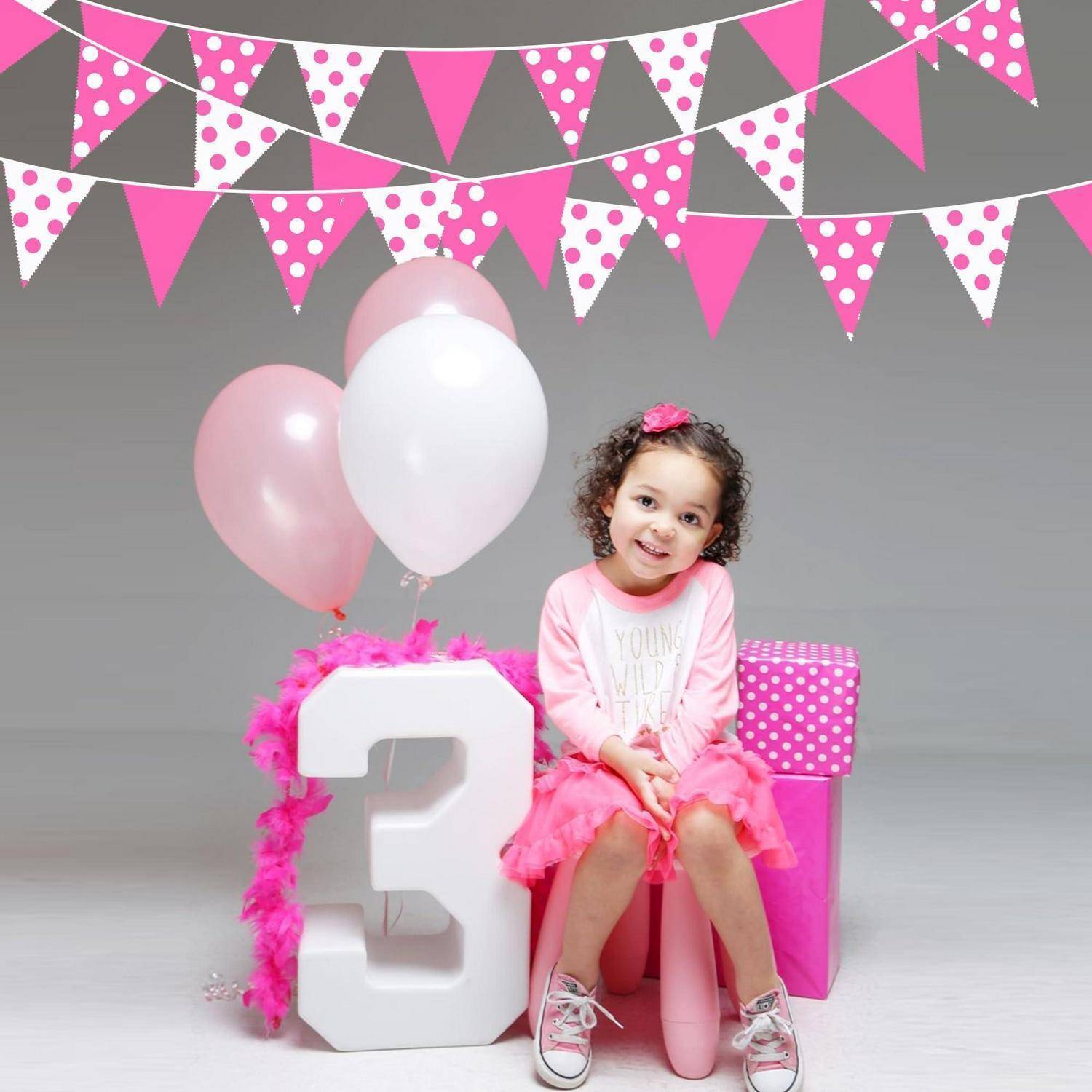 Ein kleines Mädchen in Rosa sitzt neben einer großen weißen Nummer 3 mit Ballons und Partydekoration, was auf eine Feier zum dritten Geburtstag hindeutet.