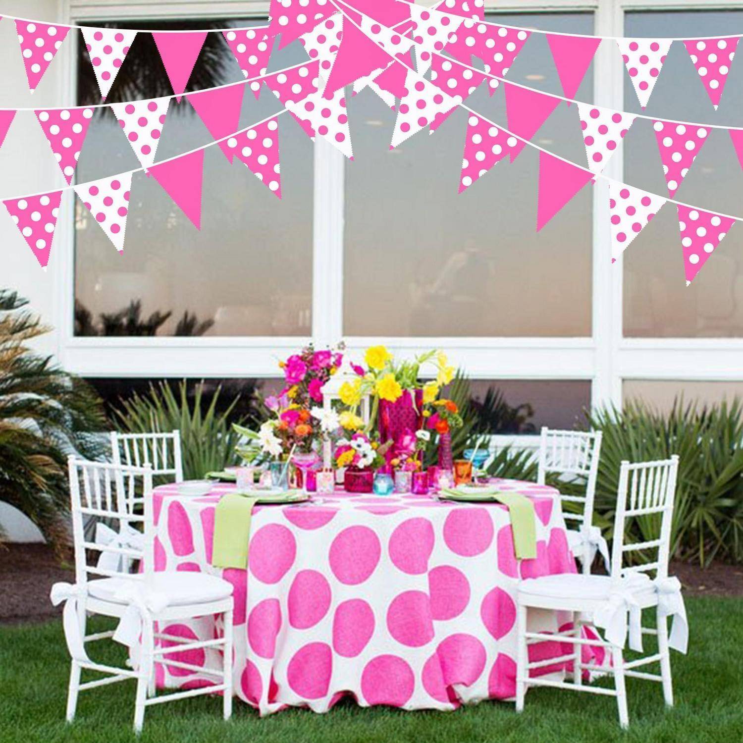 Outdoor-Partytisch mit rosa Tischdecke mit weißen Punkten, Blumenmittelstück, weißen Stühlen und rosa Wimpelgirlanden, die oben aufgehängt sind.