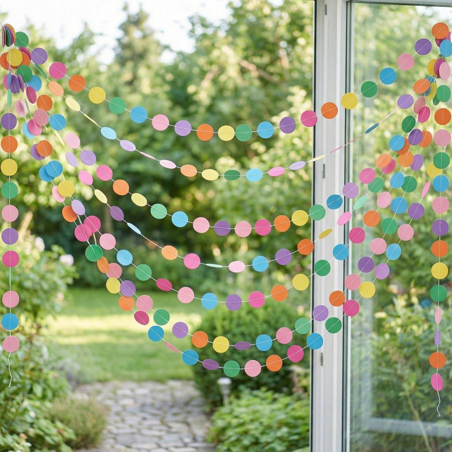 Bunte Papierpunkt-Girlanden, die über ein offenes Fenster drapiert sind und einen Blick auf einen sonnendurchfluteten Gartenweg draußen rahmen.