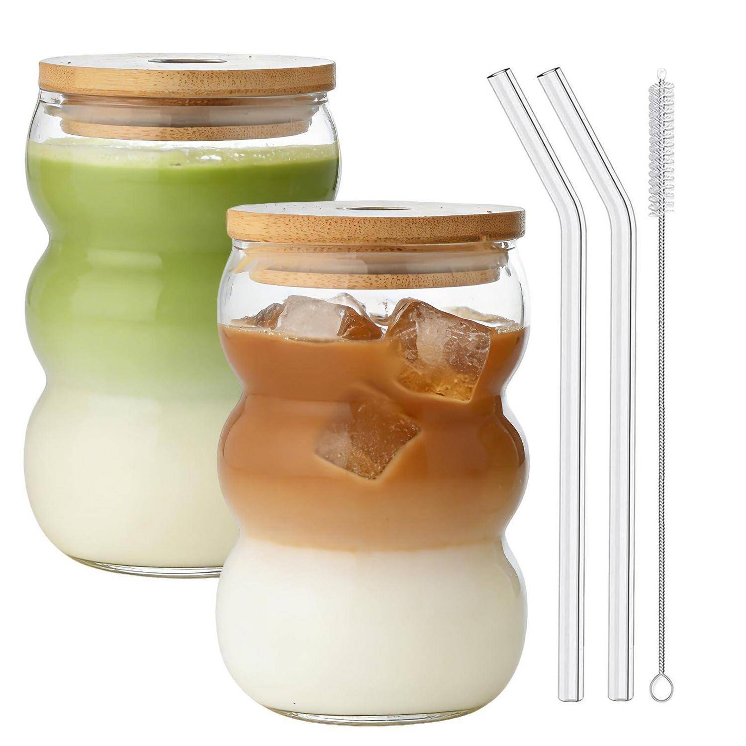 Zwei geschichtete Getränke in Glaskrügen mit Bambusdeckeln, eins grün und eins braun über Milch, zusammen mit zwei Glashalmem und einer Reinigungsbürste gezeigt.
