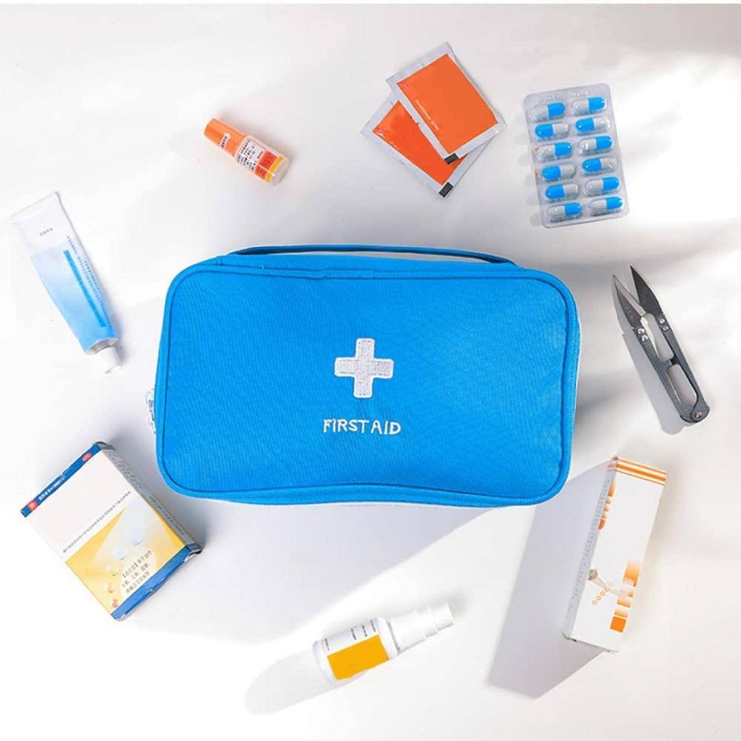 Blaukasten für Erste Hilfe mit weißem Kreuz, umgeben von Medikamentenpaketen, Pillen, Tube, Salbe und Pinzette auf weißem Hintergrund.