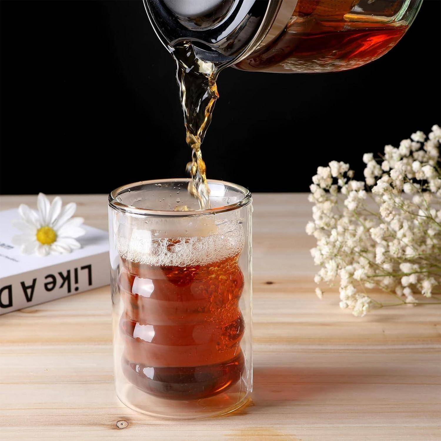 Tee wird aus einer gläsernen Teekanne in eine durchsichtige doppelwandige Glastasse auf einem Holztisch gegossen, mit Blumen in der Nähe.