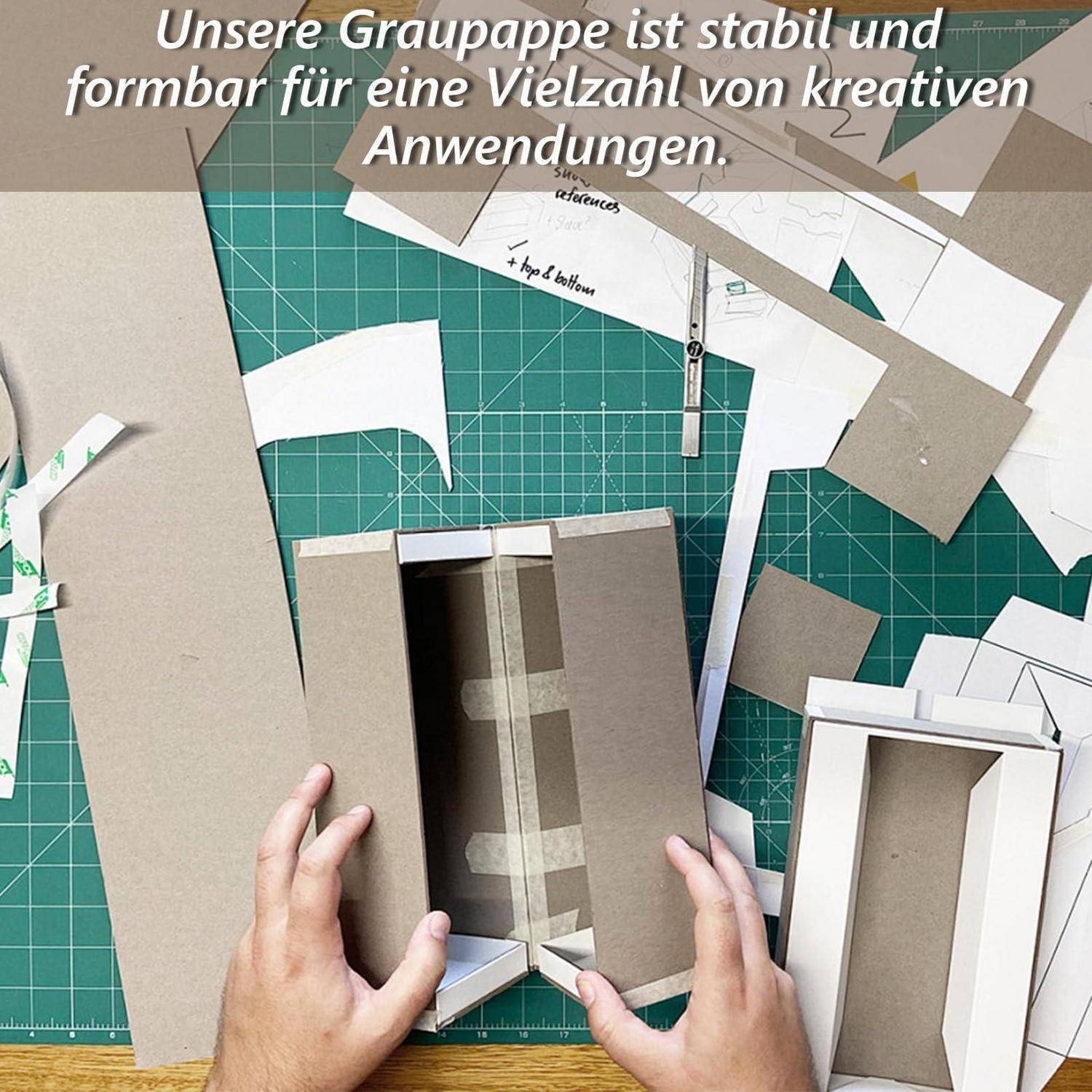 Hände falten graue Pappe zu einer Schachtel auf einer Schneidematte, umgeben von Papiervorlagen; der deutsche Text besagt, dass die Pappe stabil und vielseitig für kreative Anwendungen einsetzbar ist.