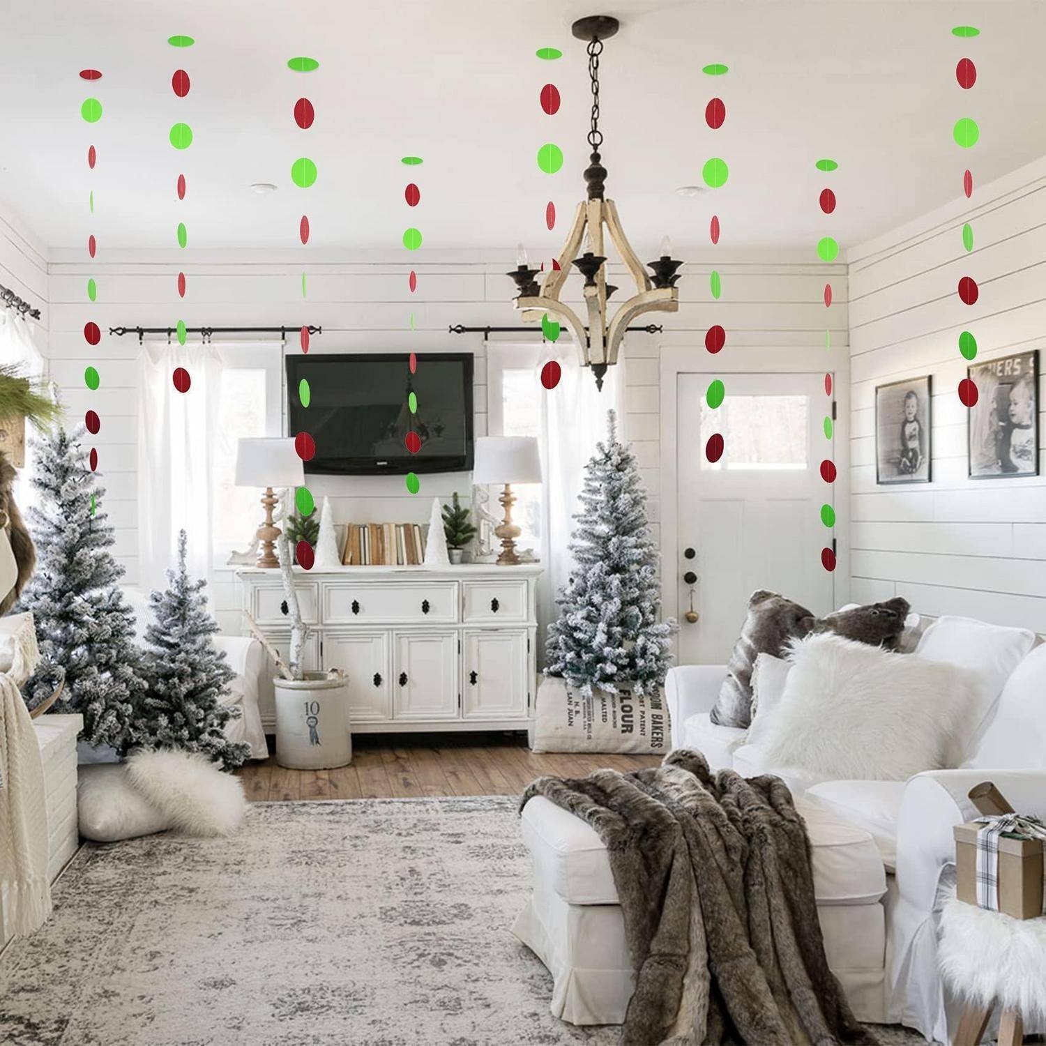 Festliches weißes Wohnzimmer mit hängenden roten und grünen Ornamentgirlanden, mit Kunstschnee bedeckten Weihnachtsbäumen und einem Sofa mit Kissen und Decken.