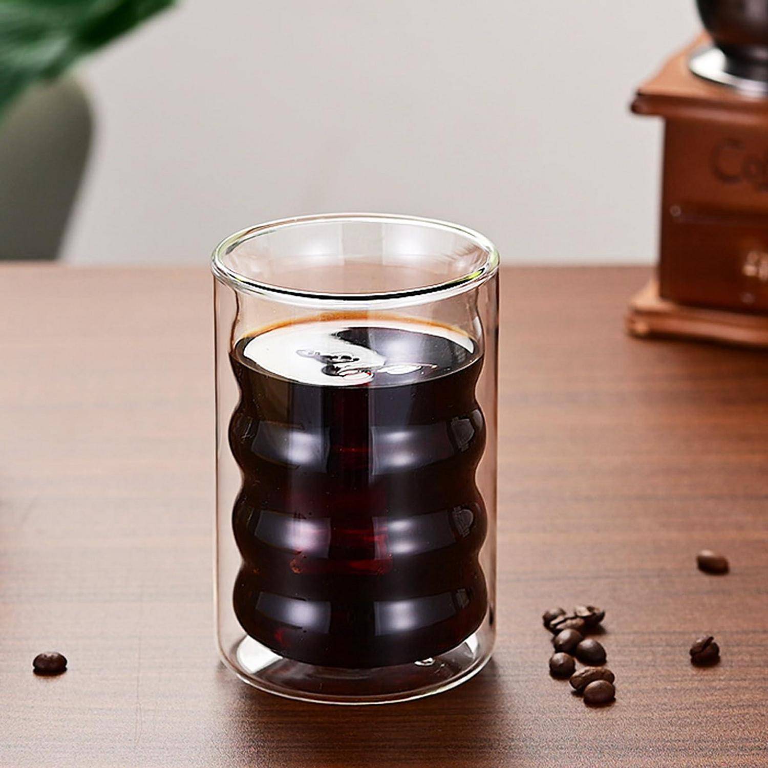 Ein klares doppelwandiges Glas gefüllt mit dunklem Kaffee sitzt auf einem Holztisch, mit verstreuten Kaffeebohnen in der Nähe.