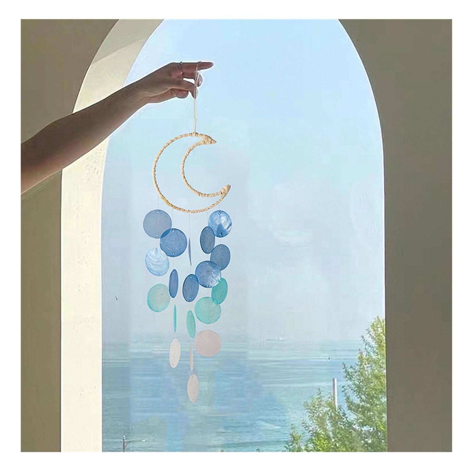 Eine Hand hält ein Wind-Chime aus Mond- und Mondlicht-Scheiben in Blau und Blaugrün in einem gewölbten Fenster mit Blick auf den Ozean.