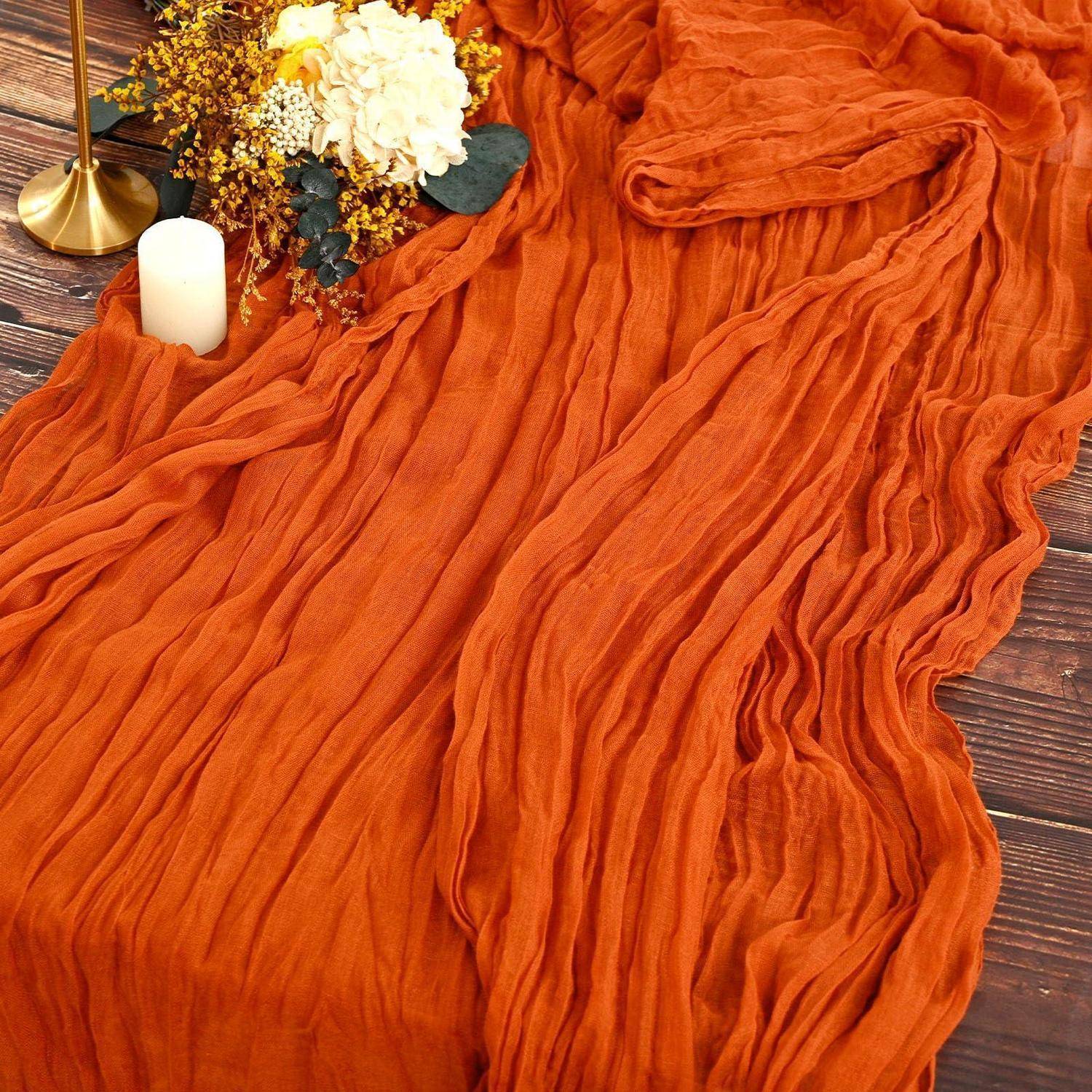 Verbrannte Orange strukturierter Stoff, der über einen Holzboden drapiert ist, neben einer Kerze, einer Goldlampe und einer herbstlichen Blumengestaltung.