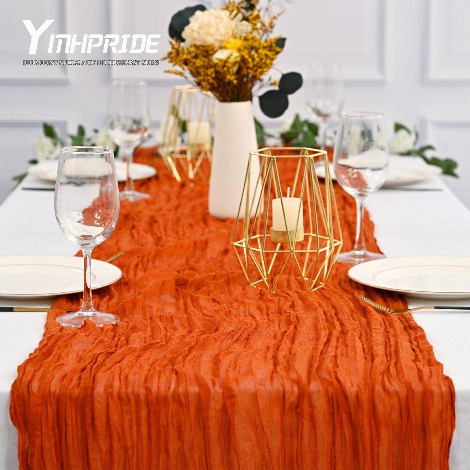 Esstisch gedeckt mit orangefarbenem strukturiertem Tischläufer, Weingläsern, weißen Tellern, Vase mit Trockenblumen und goldenem Kerzenständer als Tischdeko.