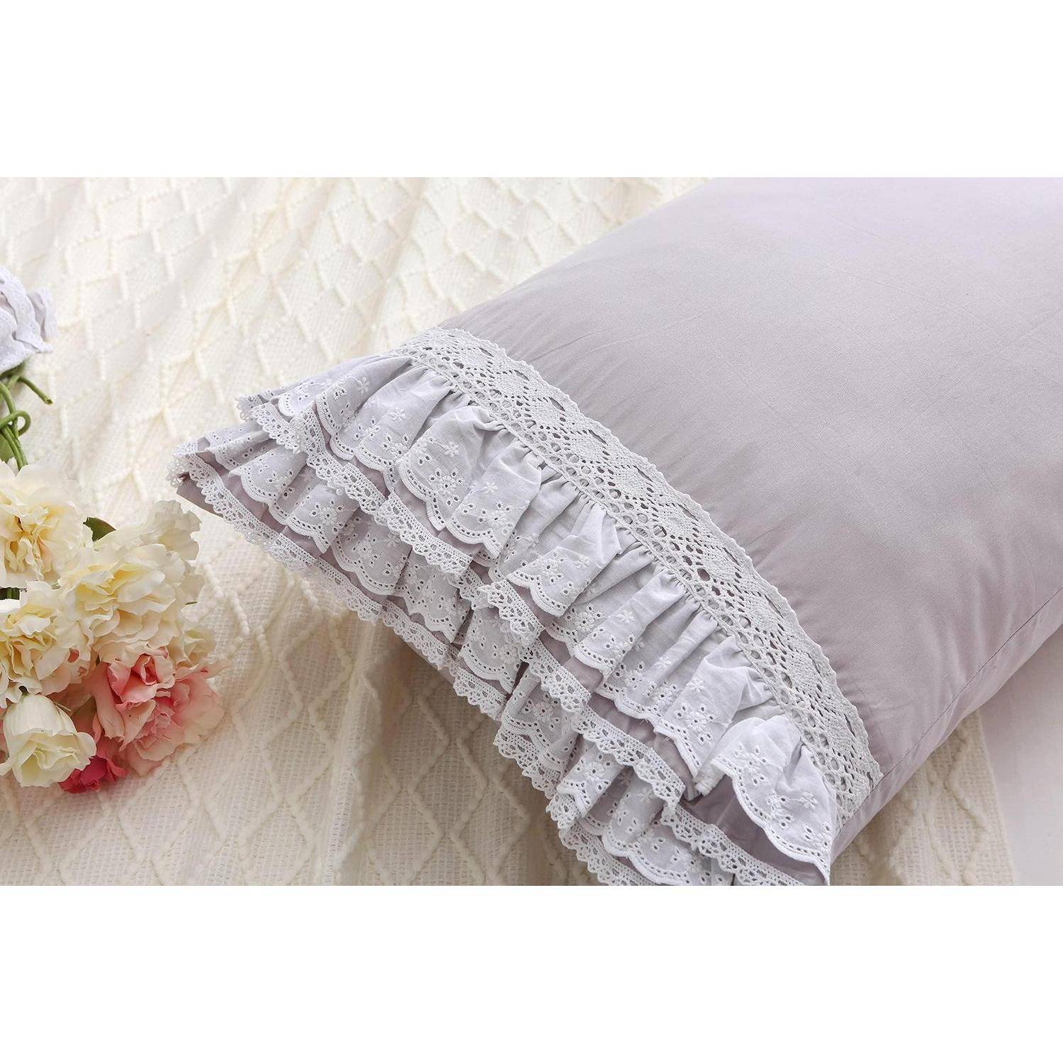 Hellblauviolettes Kissen mit geschichteter weißer Spitze und gerüschtem Saum, gezeigt neben einem Blumenstrauß auf einem Bett