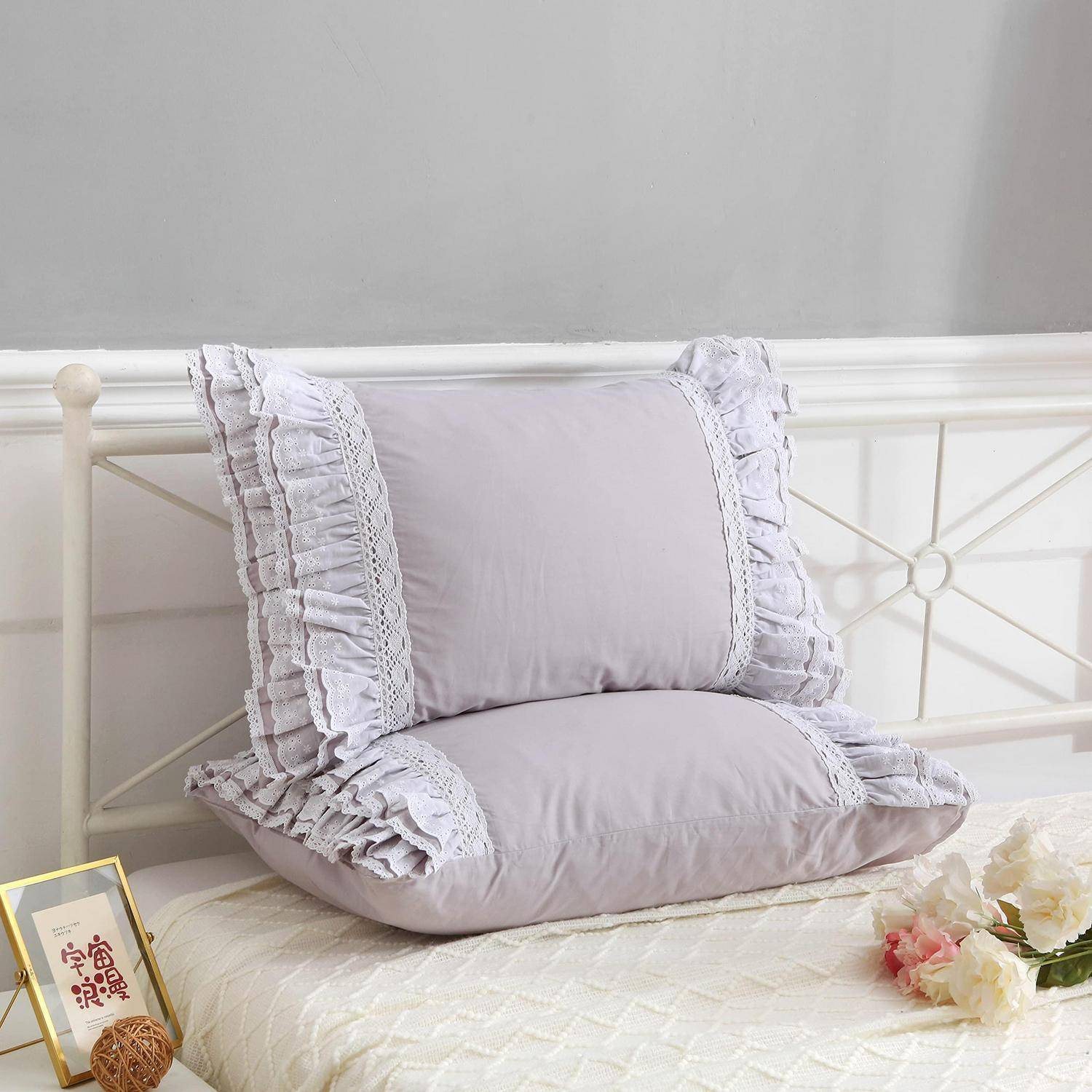 Zwei lavendelblau gerüschte Kissen, die auf einem weißen Metallbett mit einer gesteppten Decke und Blumen daneben gestapelt sind.