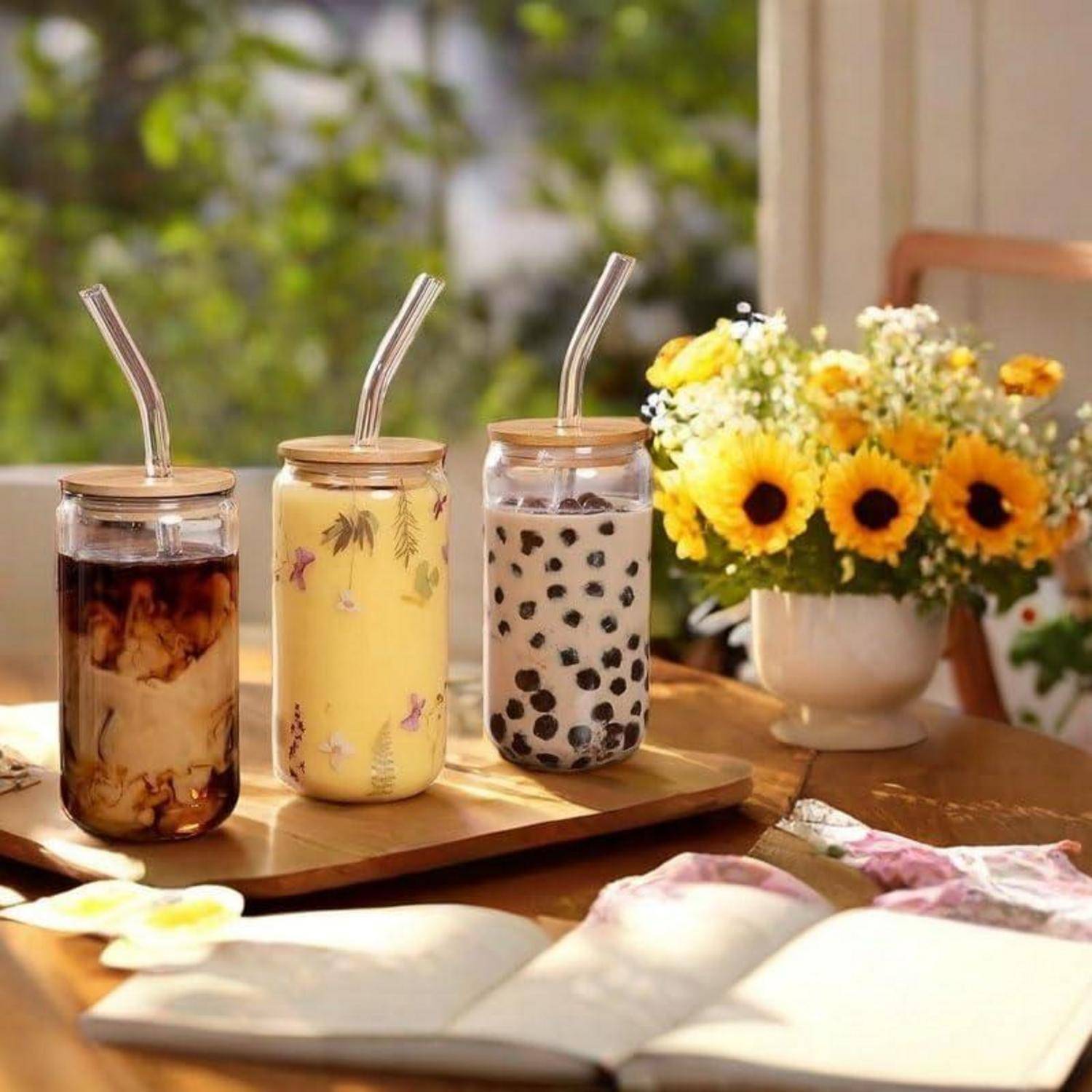 Drei gekühlte Getränke in Glasgefäßen mit Strohhalmen stehen auf einem Tablett neben einer Vase mit gelben Blumen auf einem sonnenbeschienenen Tisch.