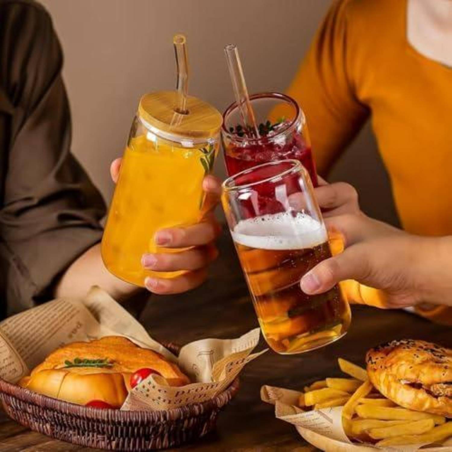Drei Personen stoßen mit Getränken über einen Tisch an, auf dem Burger, Pommes und Sandwiches stehen, was auf ein ungezwungenes Essen oder Treffen hindeutet.