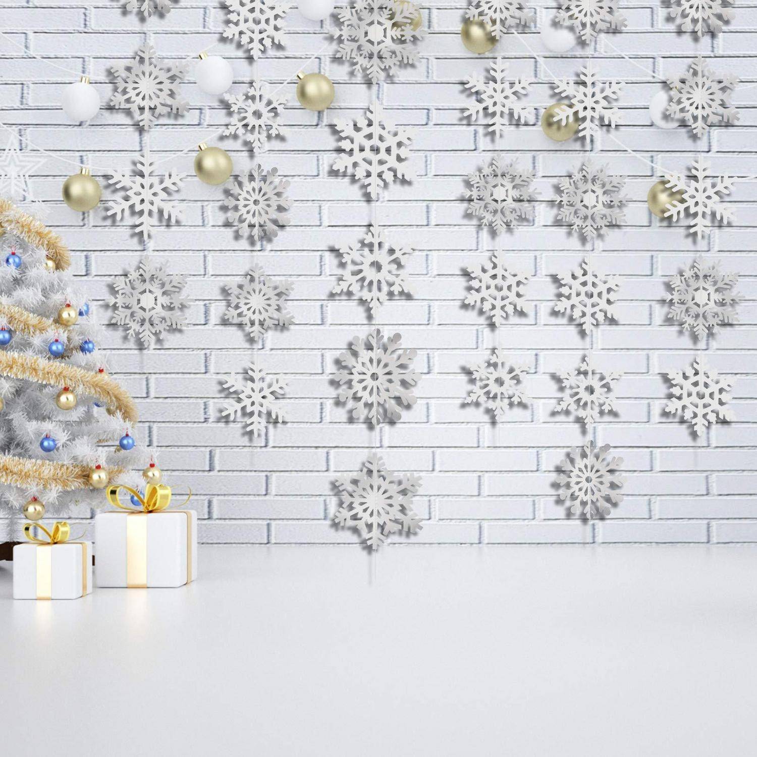 Festliche weiße Weihnachtsdeko mit einem geschmückten Baum, Geschenkboxen und hängenden Schneeflocken-Ornamenten vor einer weißen Ziegelmauer.