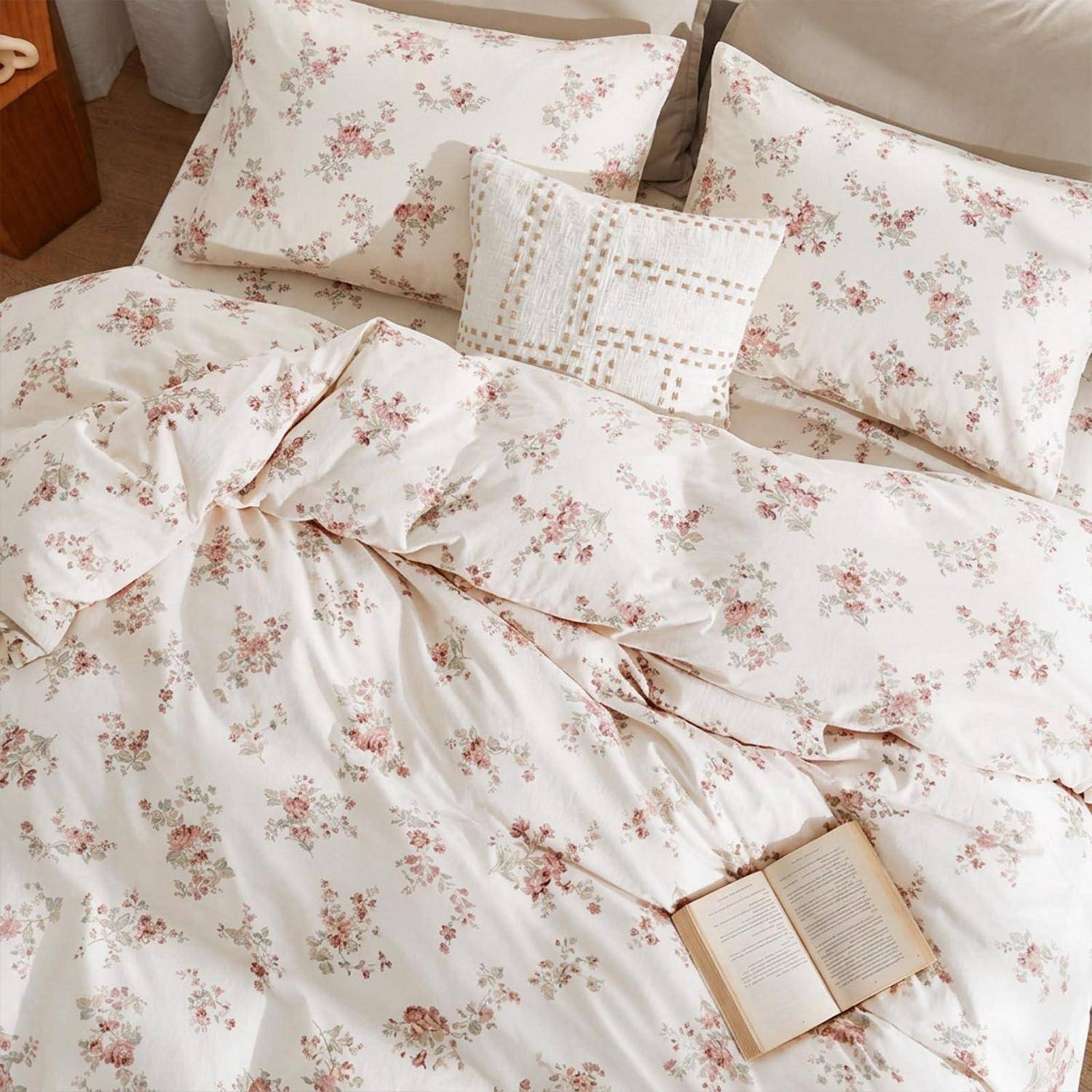 Blumige Creme-Bettwäsche mit zerknitterter Bettdecke, zwei Kissen, einem Spitzenkissen und einem aufgeschlagenem Buch auf dem Bett.