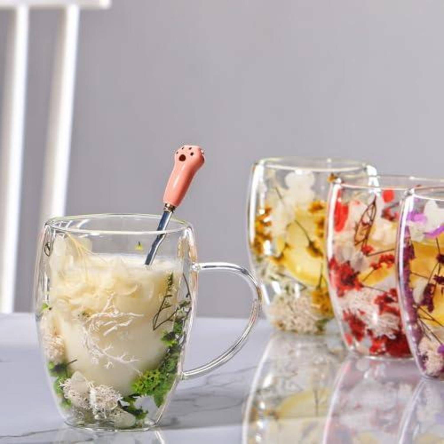 Durchsichtige Tassen mit geschichtetem Dessert oder Tee gefüllt, eine mit einem Löffel mit korallenrotem Griff, und drei bunte Frucht- und Blütengetränke im Hintergrund.