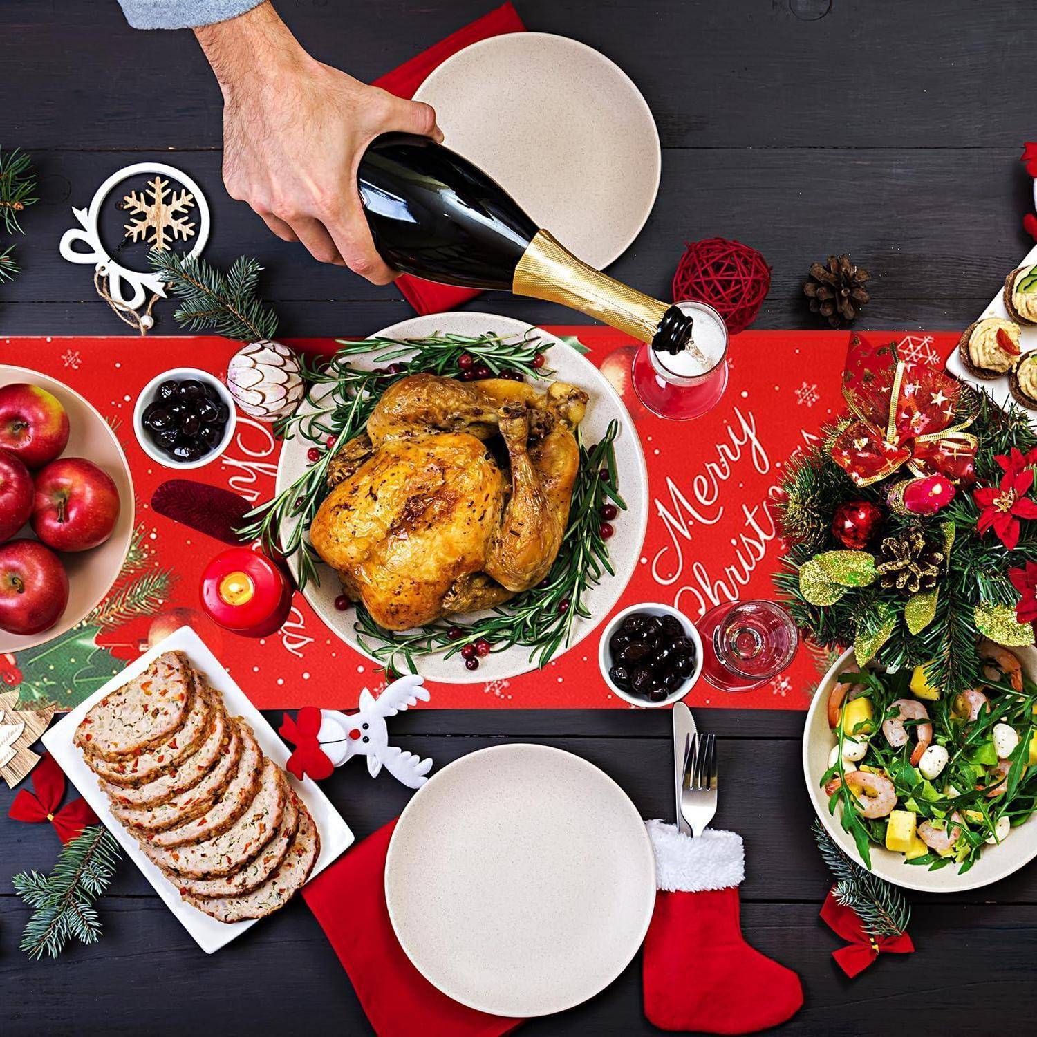 Von oben fotografierter Weihnachtstisch mit gebratenem Truthahn, geschnittenem Brot, Salat, Äpfeln und einer Hand, die Champagner über einen festlichen „Frohe Weihnachten"-Tischläufer gießt.