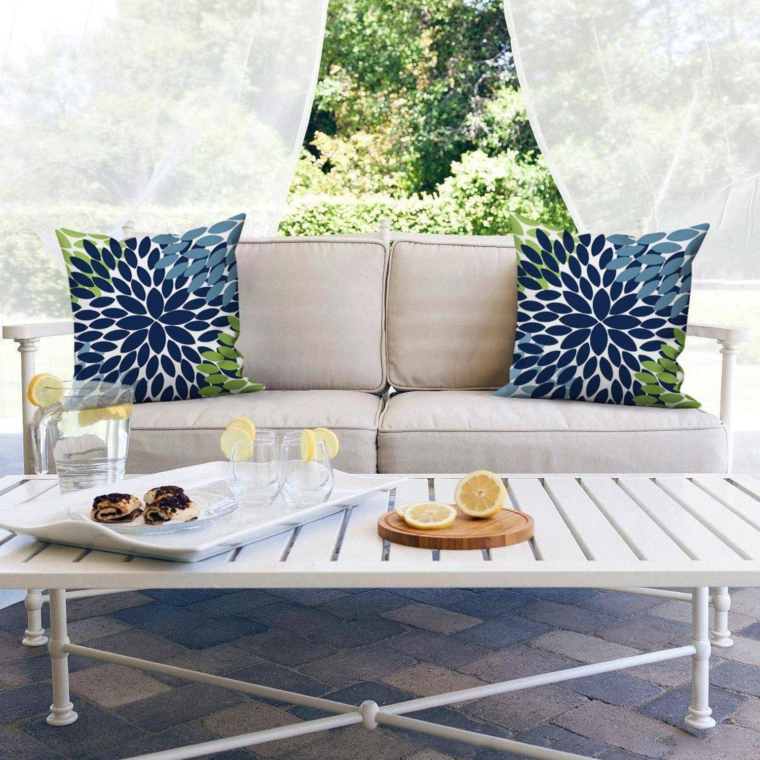 Terrassenbereich mit beigem Sofa, blau gemusterten Kissen, weißem Couchtisch mit Zitronen und Snacks sowie durchsichtigen Vorhängen, die das Grün einrahmen.