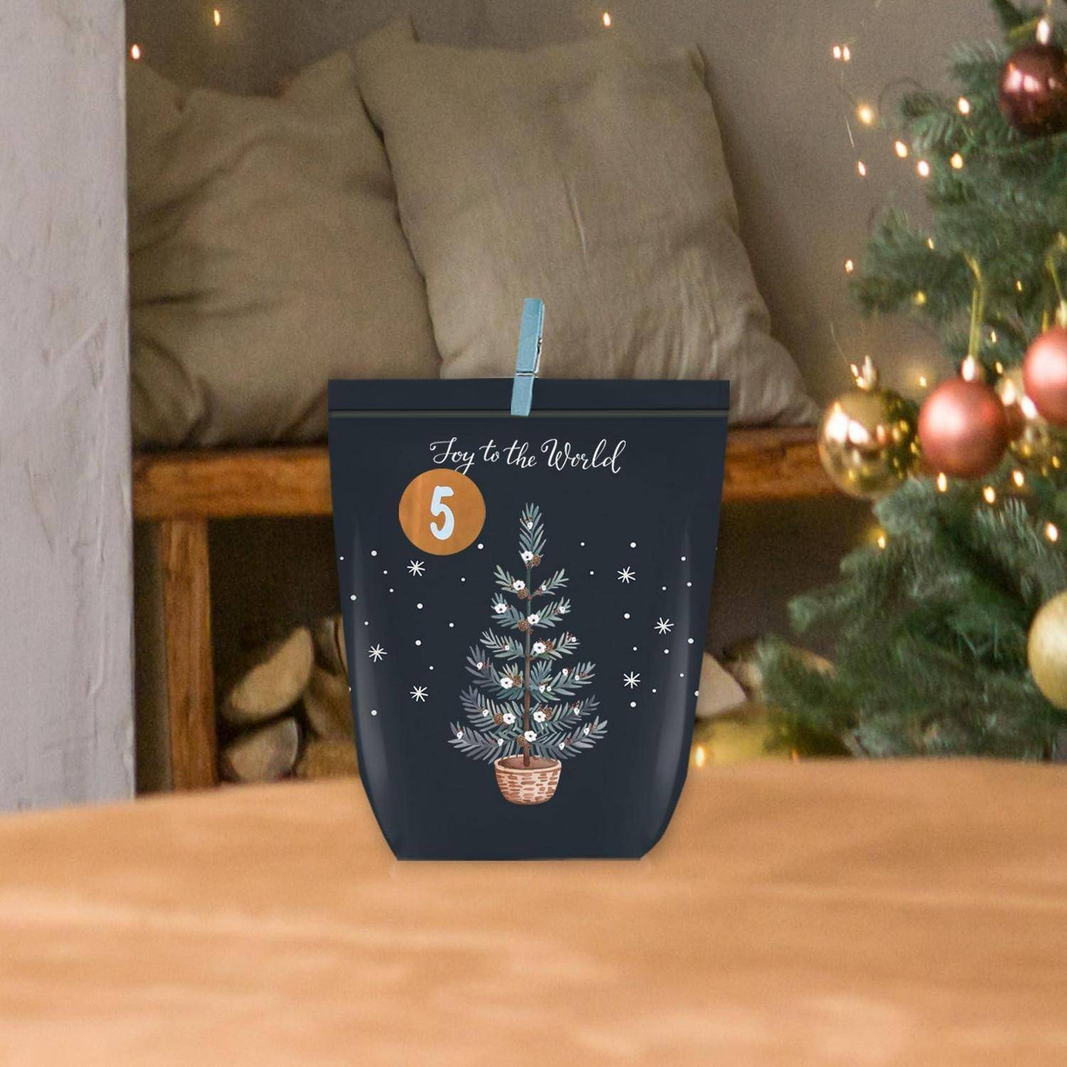 Geschenktasche für den Urlaub mit einer Weihnachtsbaumillustration und dem Text „Freude der Welt", neben einem geschmückten Baum und gemütlichem Stuhl ausgestellt.