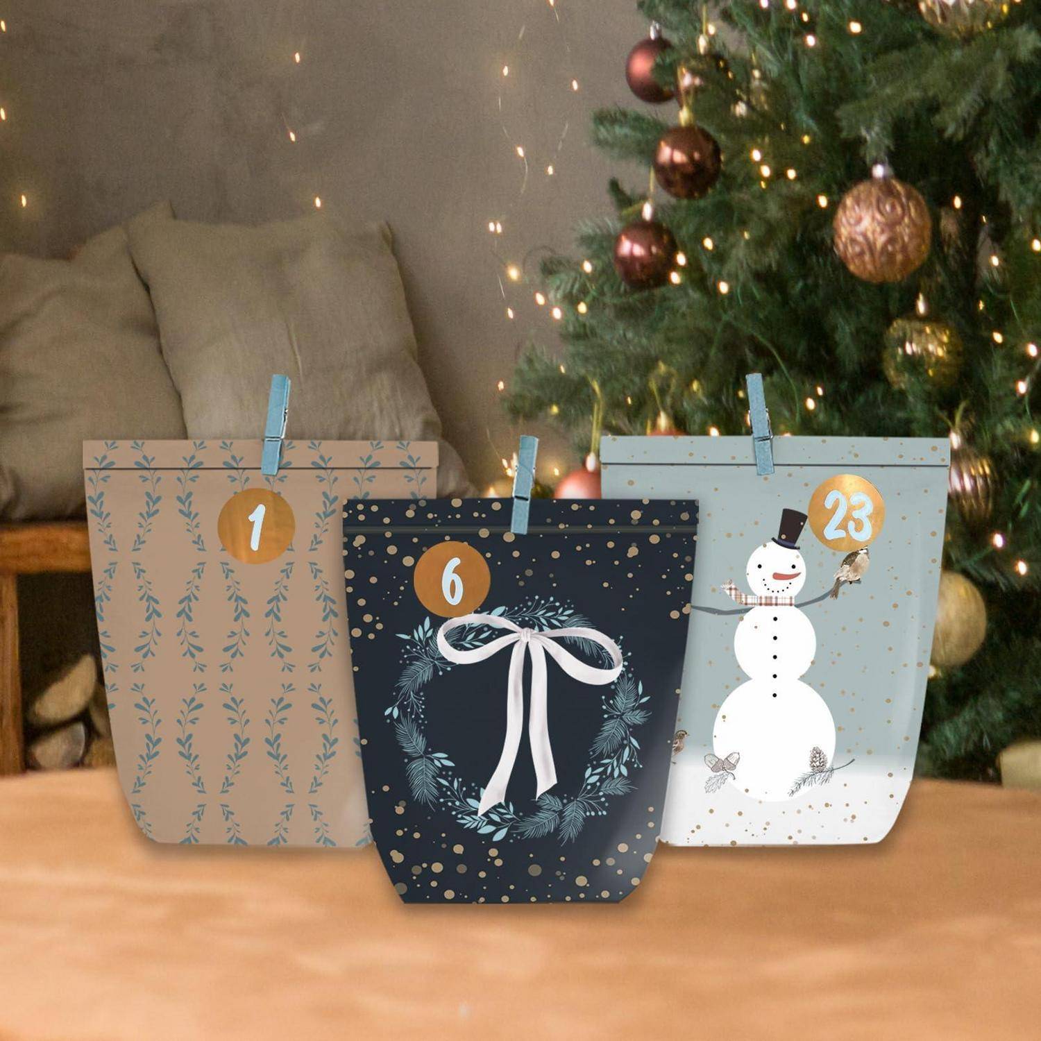 Drei festliche Papier-Adventskalender-Tüten mit den Nummern 1, 6 und 23 stehen auf einem Tisch vor einem geschmückten Weihnachtsbaum.