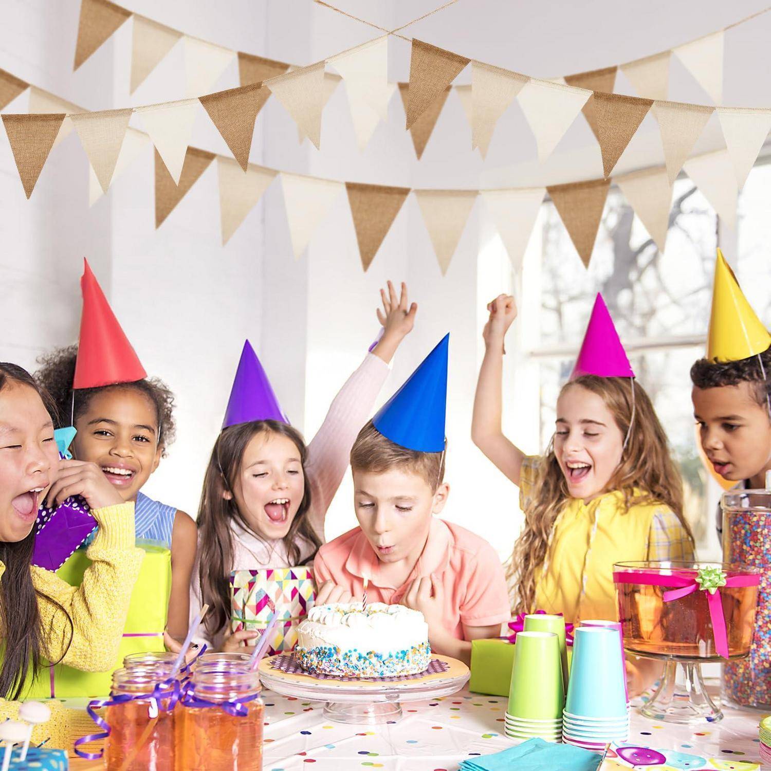 Kinder in farbenfrohen Partyhüten feiern um einen Geburtstagskuchen herum und jubeln an einem dekorierten Partytisch mit Bechern und Leckereien.