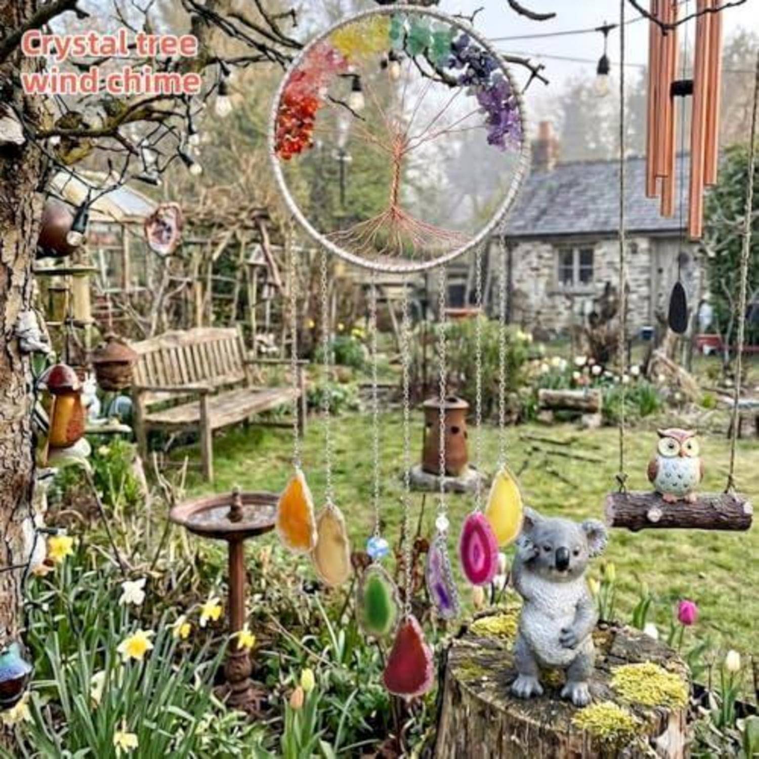 Gartenszene mit einem kristallinen Windspiel in Baumform, das über einem moosigen Baumstumpf hängt, mit einer Koala-Figur, einer Eulenornament, einer Bank, Blumen und Vogelfutterern.